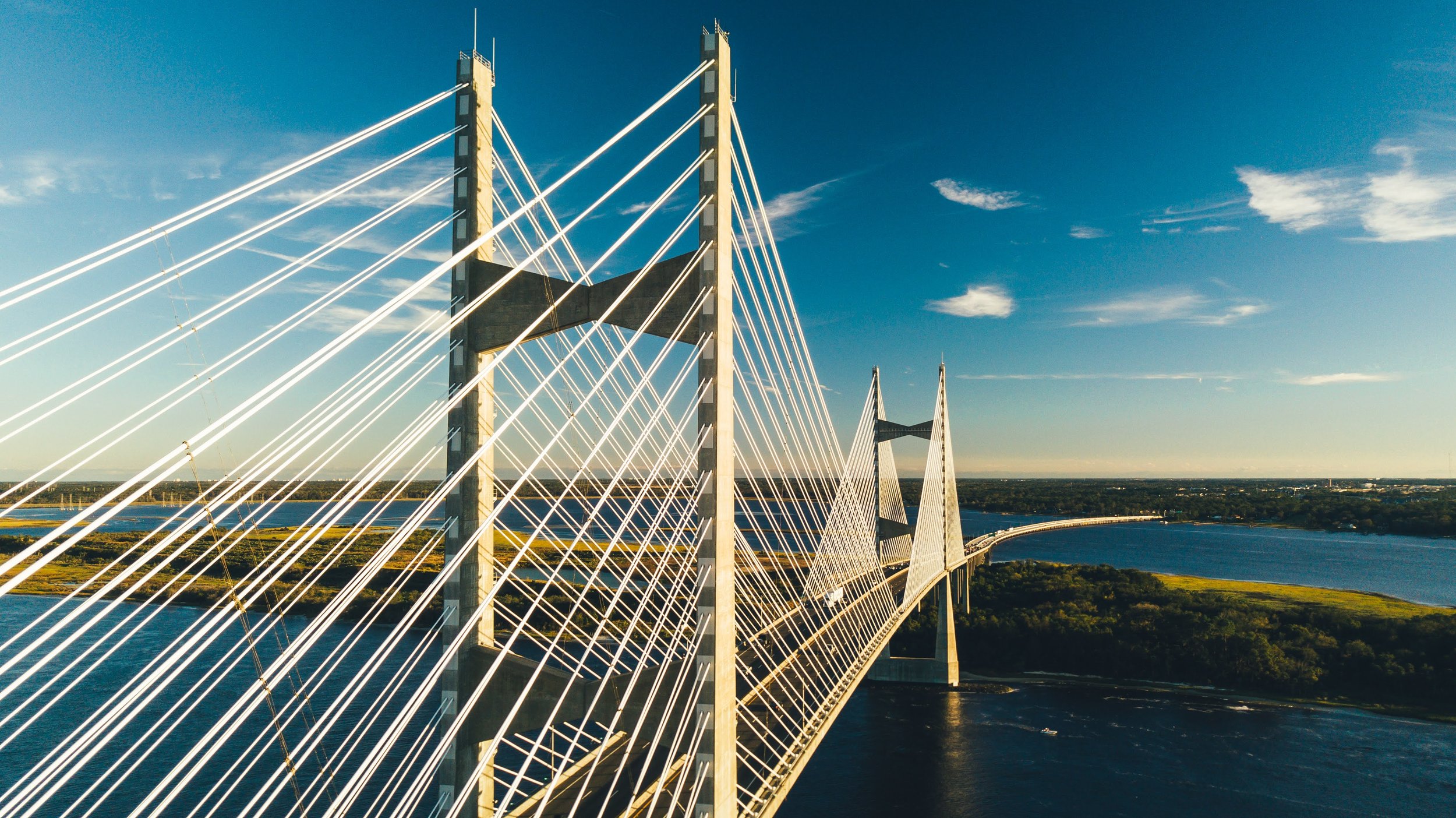 A modern cable-stayed bridge with tall pylons and white cables spanning over a wide river at sunset.