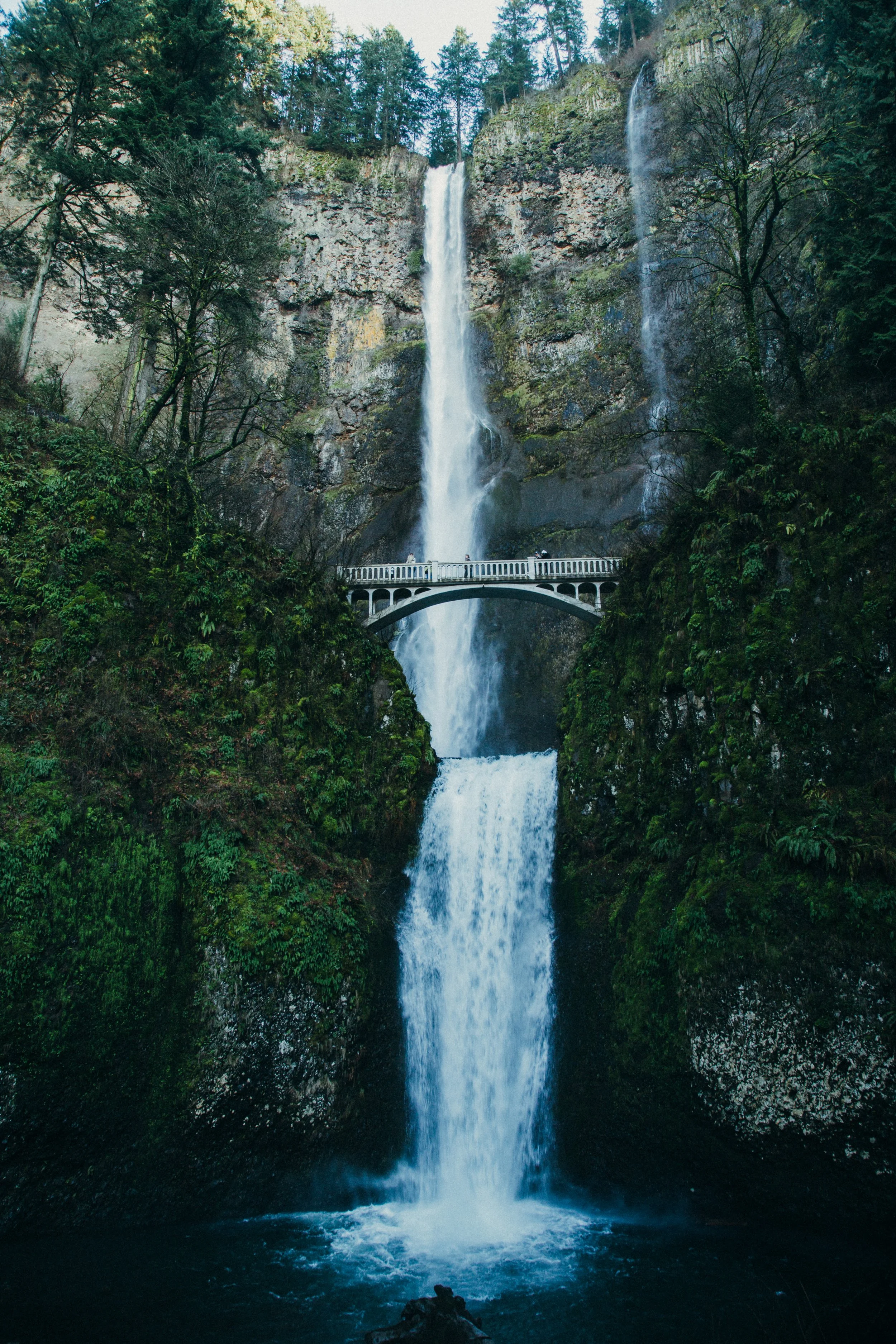 A tall multi-tiered waterfall flowing over a cliff with lush green trees surrounding it, a white bridge crossing the lower part of the waterfall.