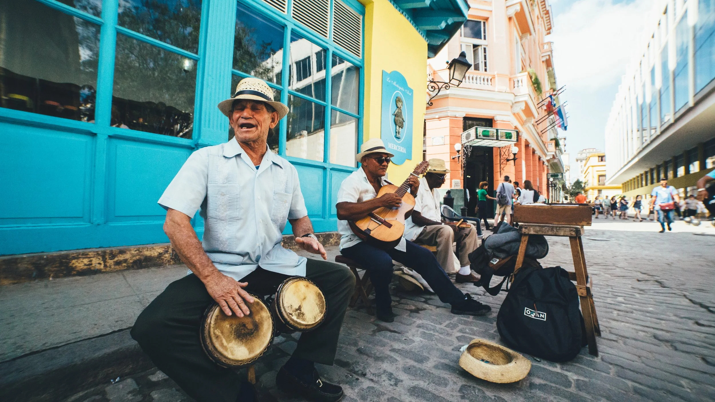 Street musicians performing on a cobblestone sidewalk, including a man playing bongos and another playing guitar, with colorful buildings and pedestrians in the background.