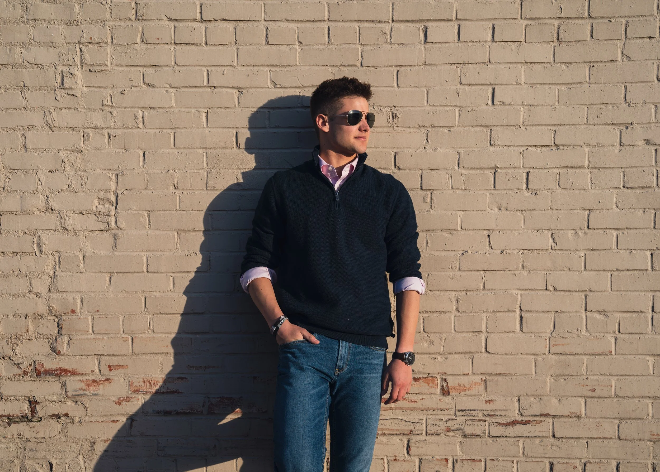 Young man wearing sunglasses, a pink collared shirt, navy sweater, jeans, and a watch, standing against a beige brick wall with sunlight casting a shadow.