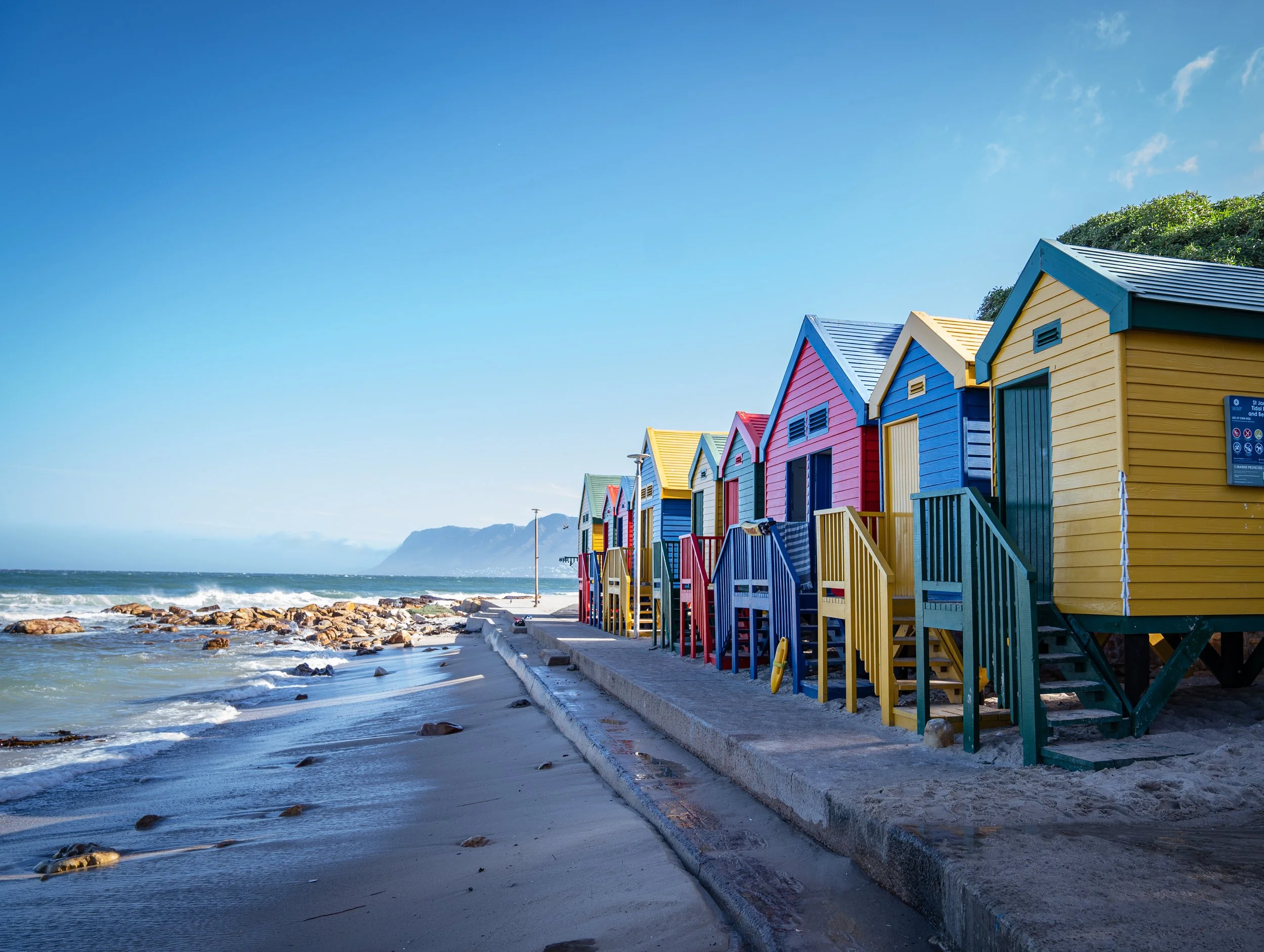 Colorful beach huts lined up along a sandy shoreline with ocean waves and distant mountains under a clear blue sky.