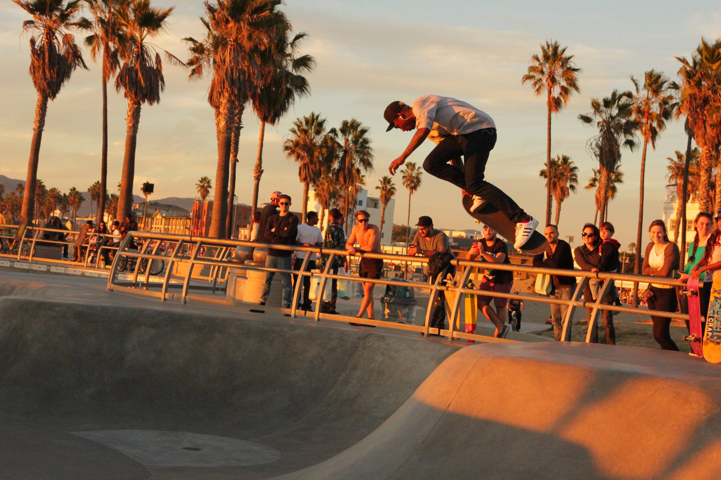A skateboarder flips his board while in mid-air at a skate park during sunset, with onlookers and palm trees in the background.