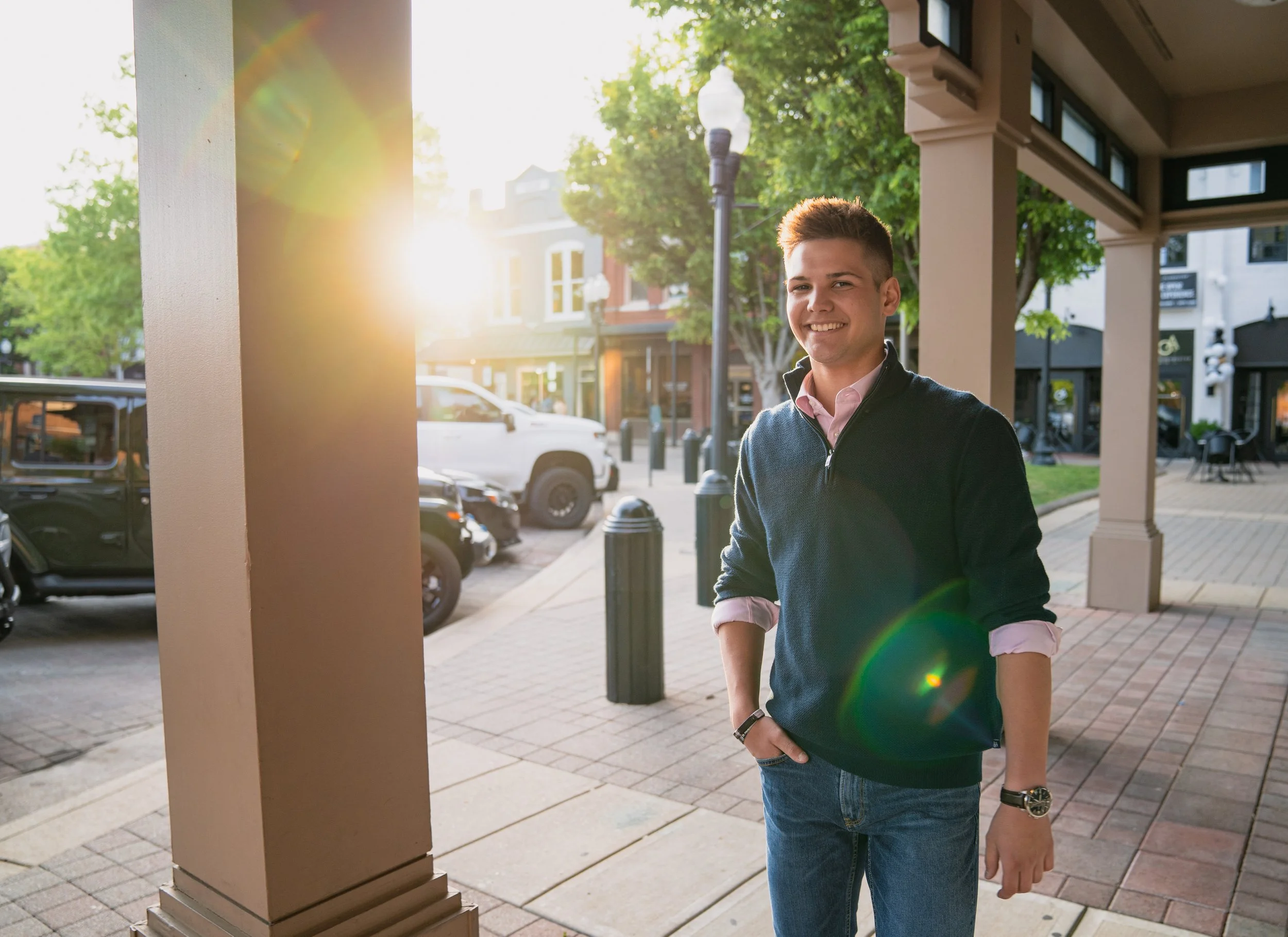 A young man with short hair, wearing a dark sweater over a light pink shirt, and jeans, standing outdoors on a city sidewalk in the late afternoon, smiling at the camera with sunlight and a rainbow lens flare in the background.