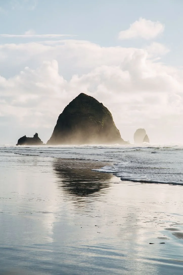 A scenic view of large rock formations rising from the ocean near a beach, with waves gently lapping the shore and a partly cloudy sky overhead.