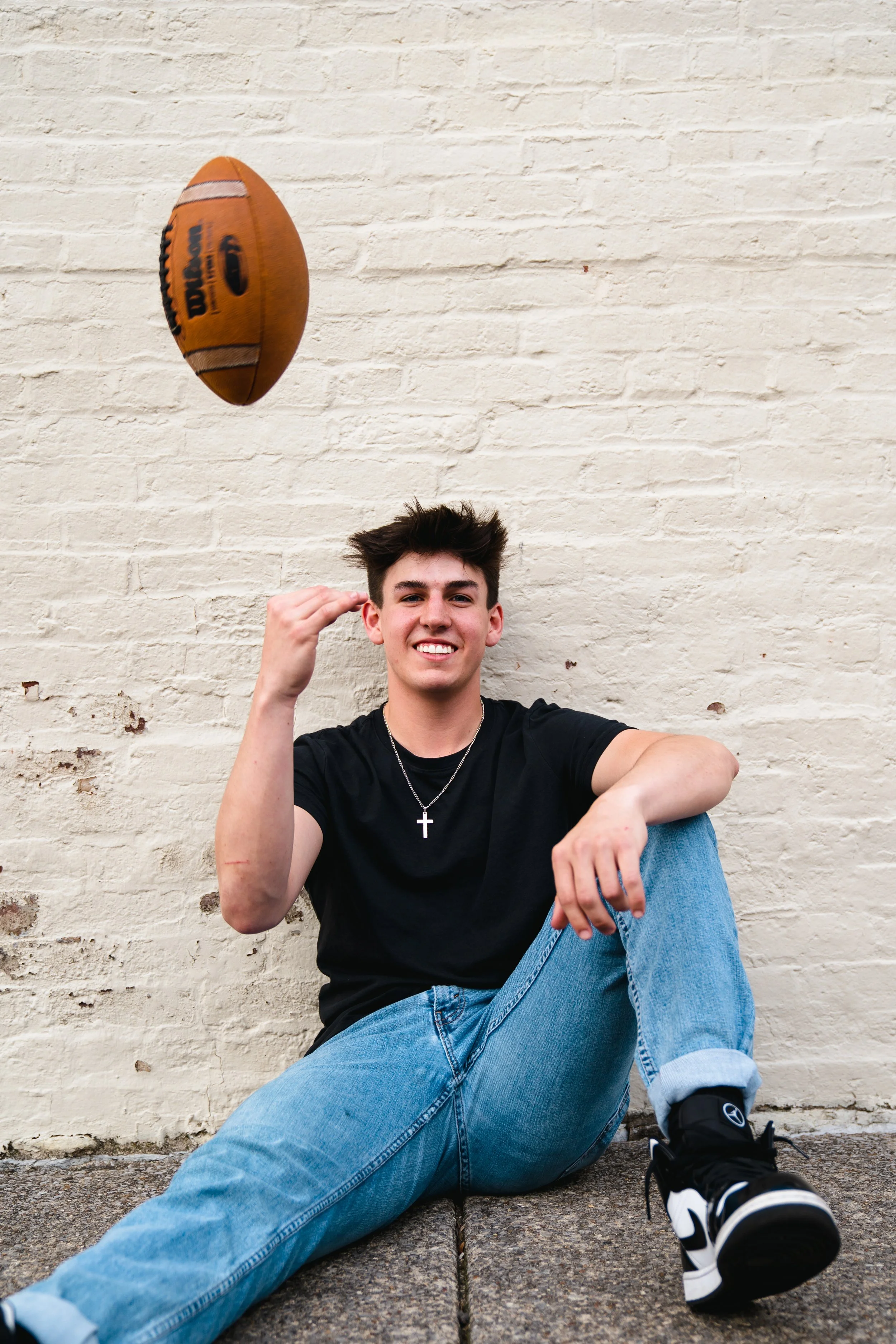 A young man sitting on the sidewalk against a white brick wall, smiling, with a football spinning in the air above him.