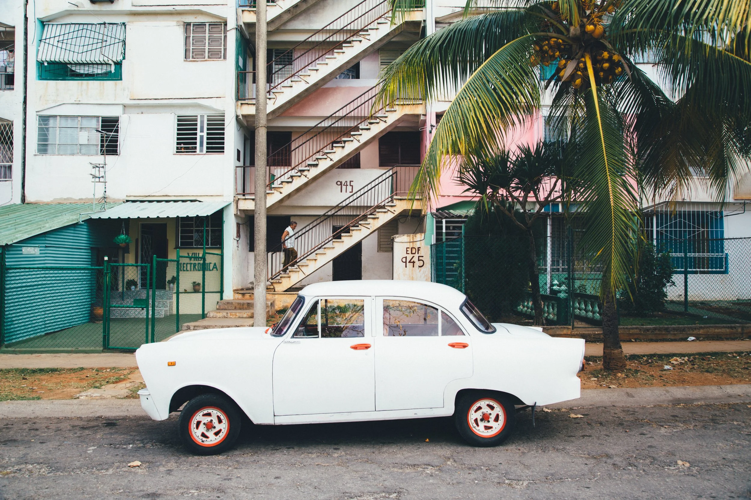 A vintage white car parked in front of a multi-story residential building with external staircase, tropical palm trees, and a man walking on the stairs.