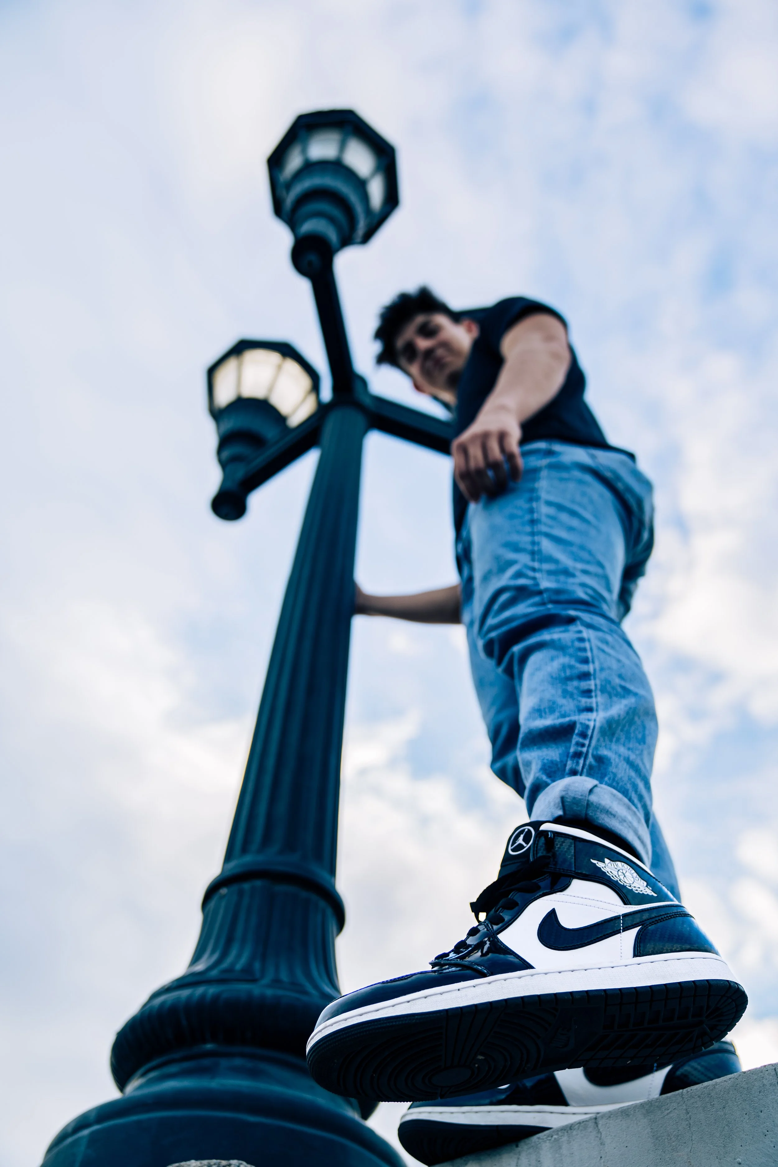Young man in jeans and black T-shirt standing next to a black street lamp, shot from below against a cloudy sky.