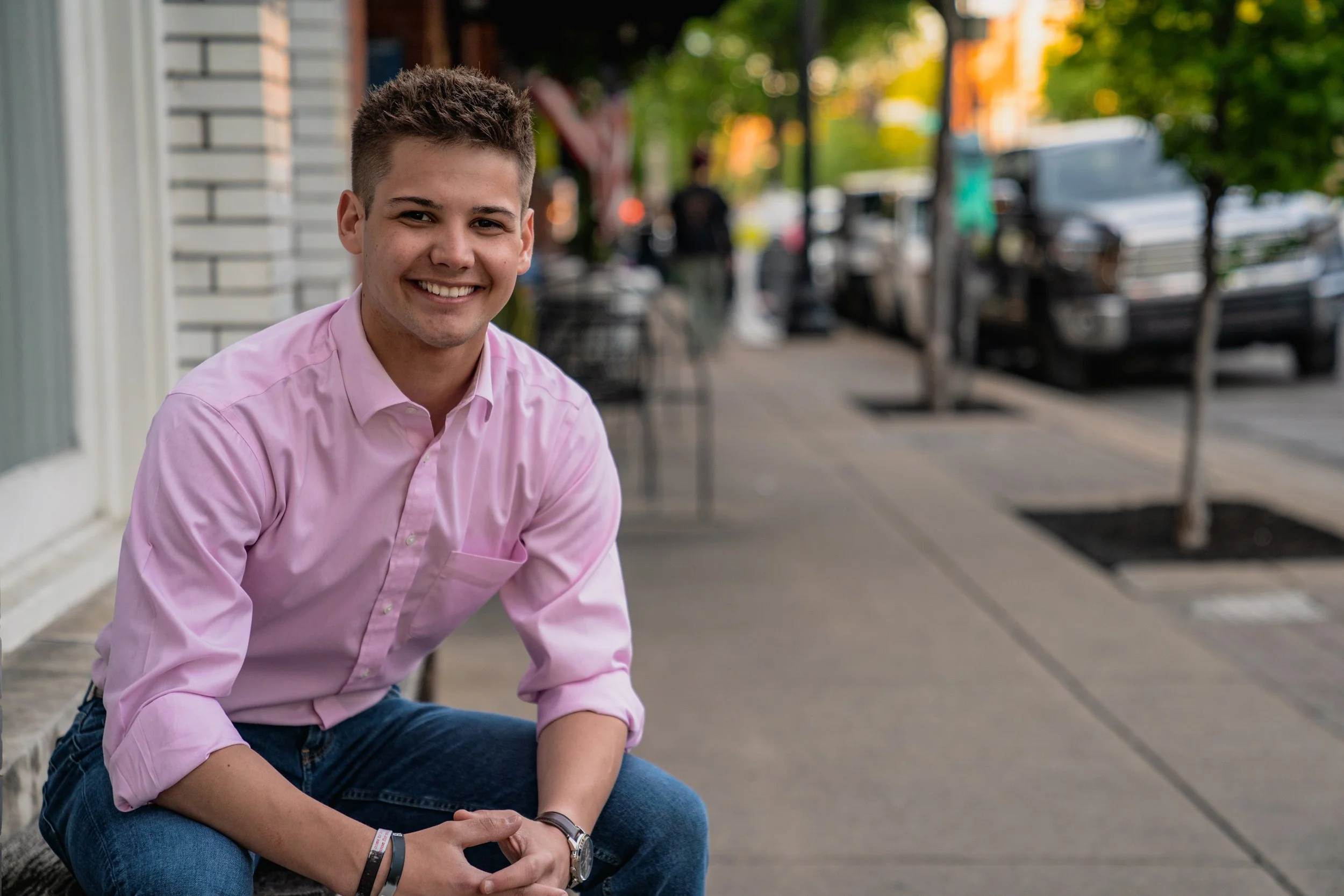 A young man sitting on a curb outside on a city sidewalk, smiling at the camera, wearing a pink button-up shirt and blue jeans.