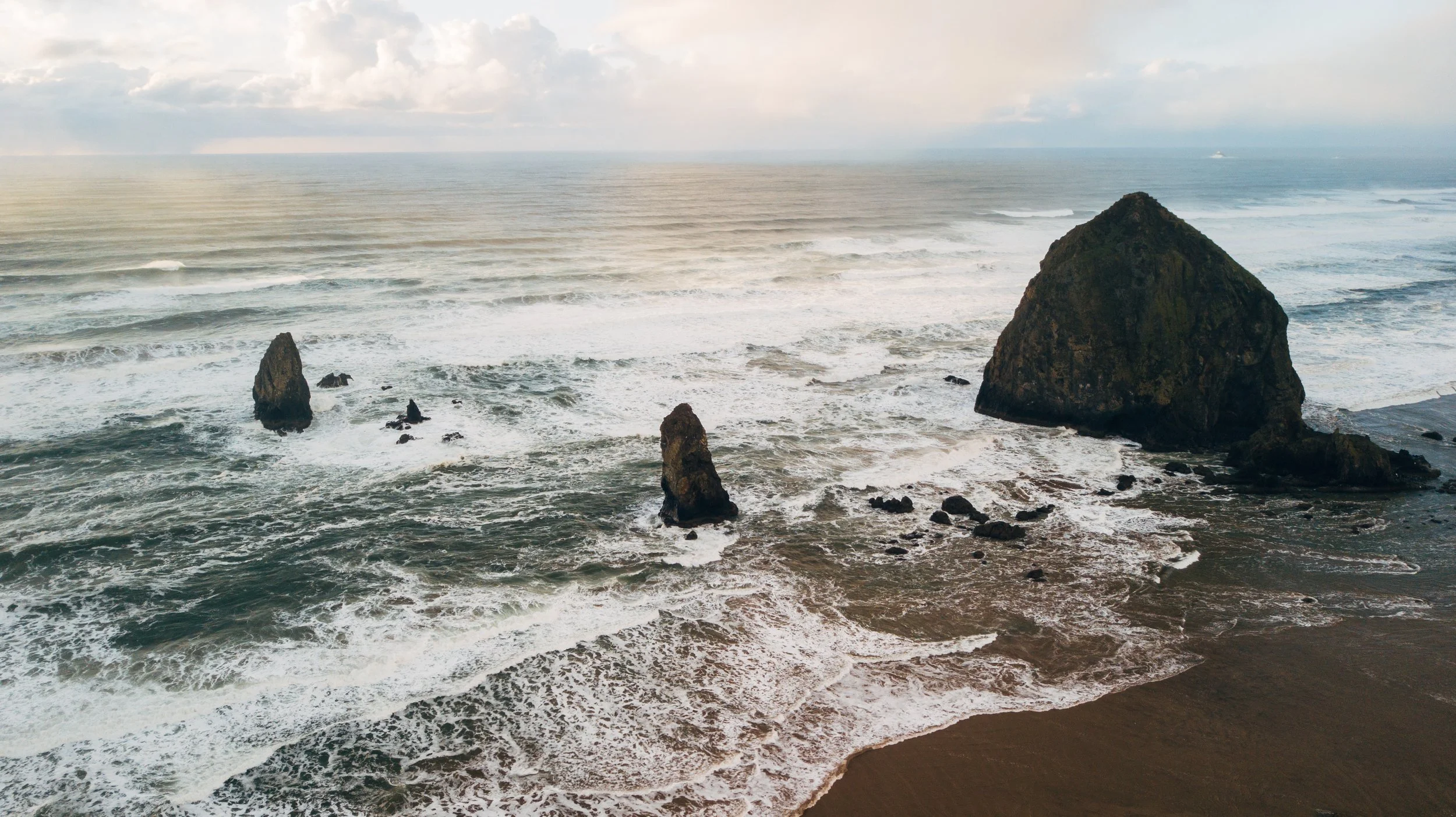 Aerial view of a rugged coastline with large rock formations protruding from the ocean near a sandy beach, under a cloudy sky.
