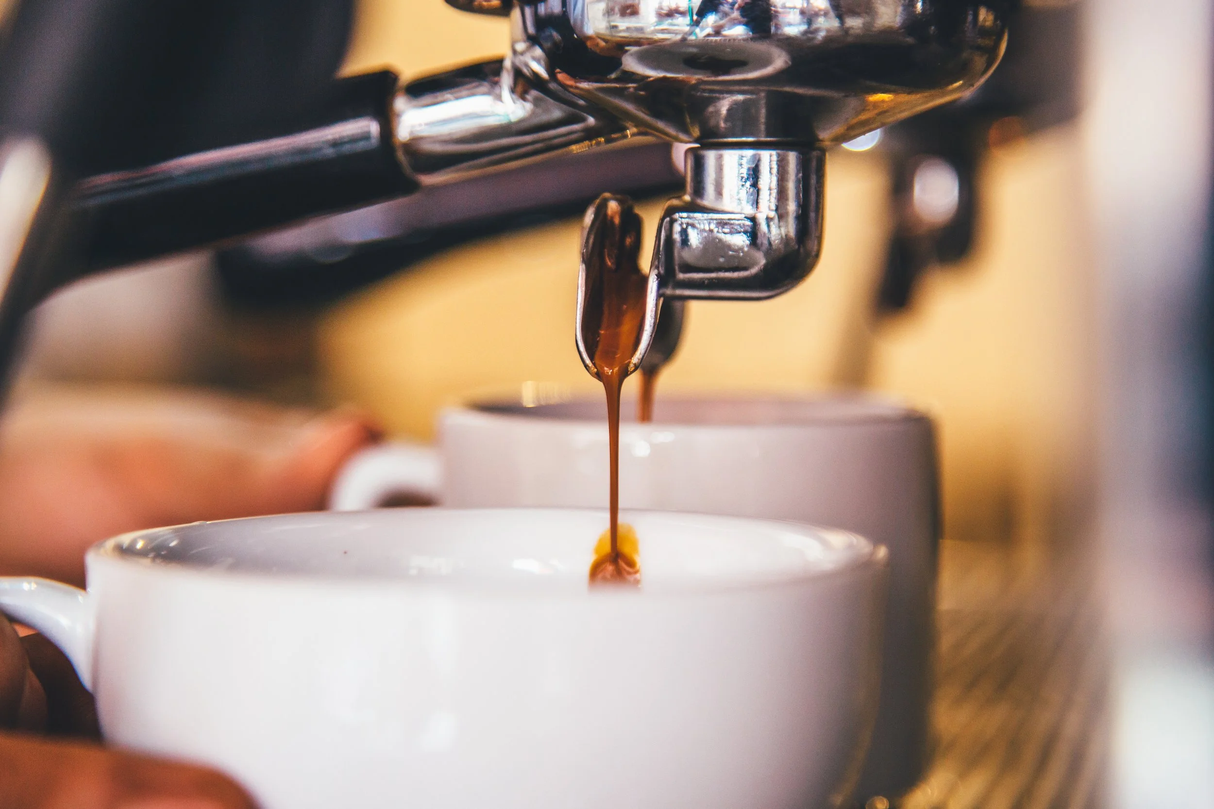 Close-up of an espresso machine pouring dark coffee into two white cups, with a person's hand holding the cup on the left side.