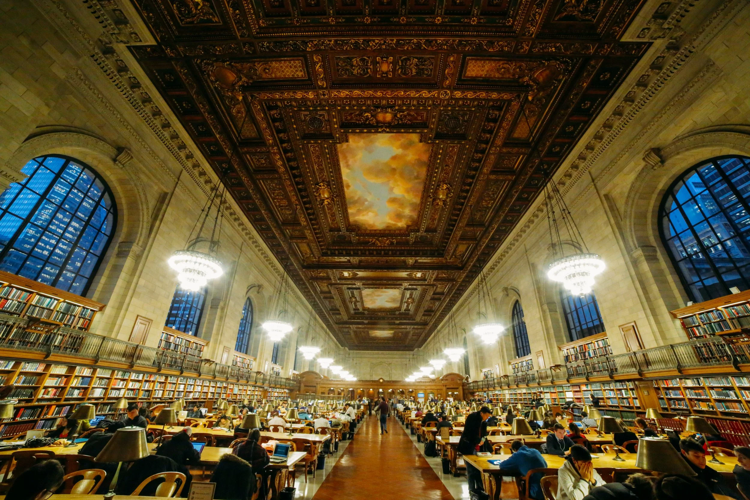 Interior of a grand historic library with a high ornate ceiling, large arched windows, tall bookshelves, and people studying at long wooden tables.