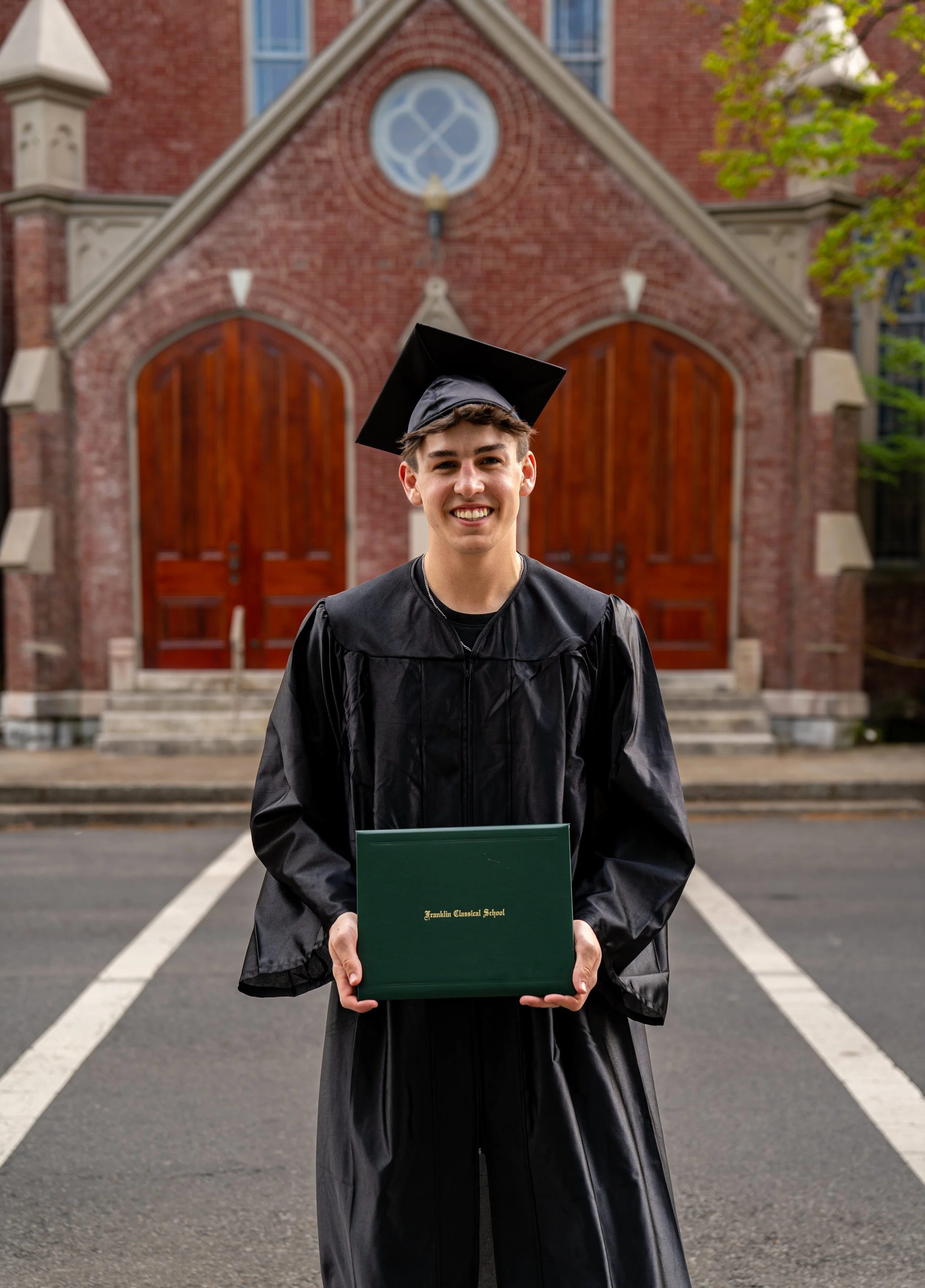 A young man in a black graduation cap and gown holding a diploma standing in front of a brick church with large wooden doors.
