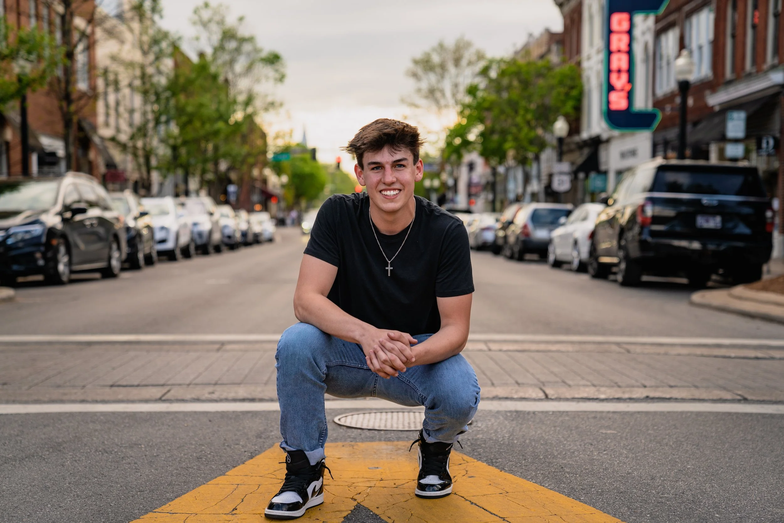A young man in a black t-shirt, blue jeans, and black and white sneakers squats on a yellow crosswalk on a city street, smiling at the camera with cars and buildings in the background.