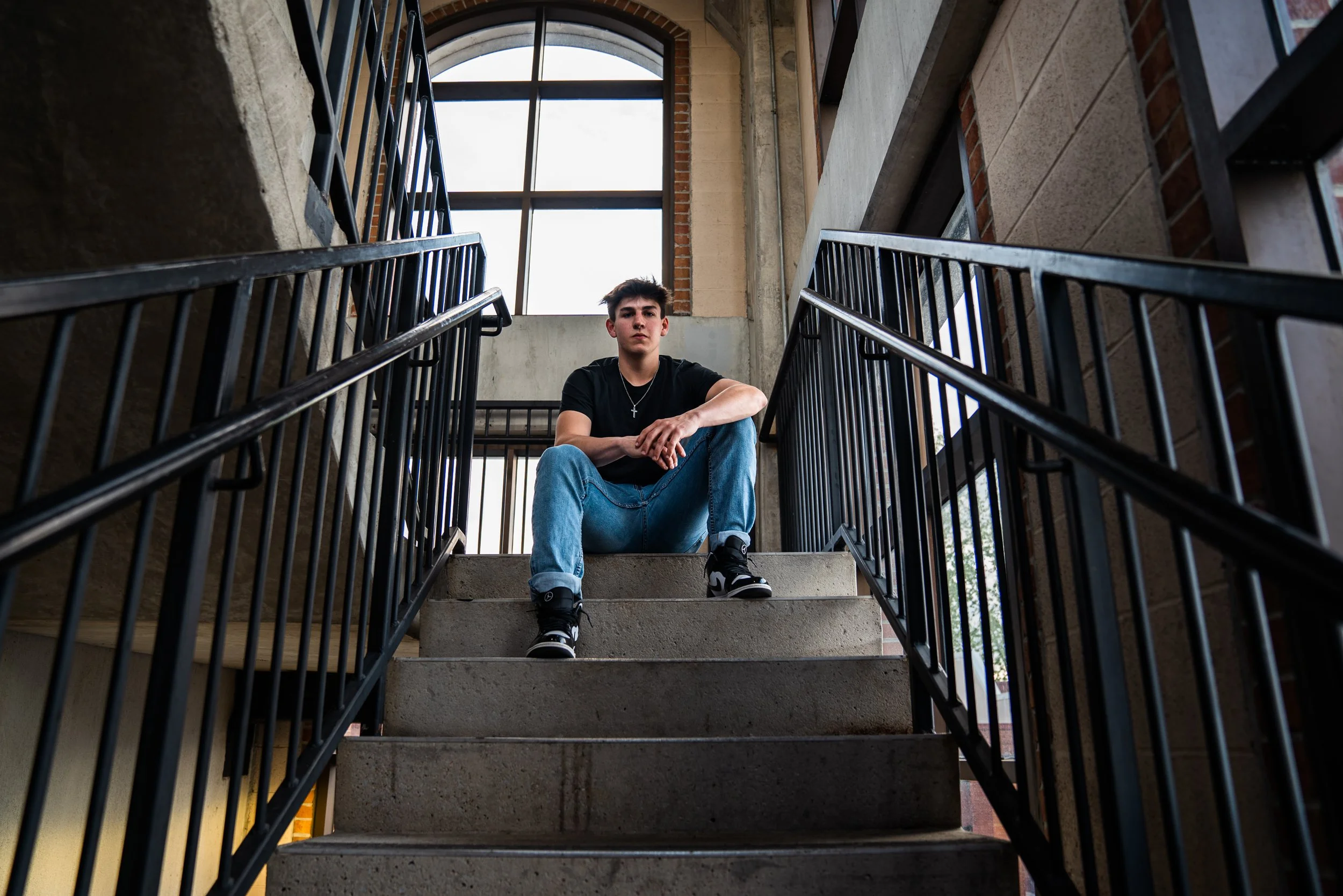 A young man with dark hair wearing a black t-shirt, blue jeans, and black sneakers sitting on concrete stairs inside a building with large windows and black metal railings.