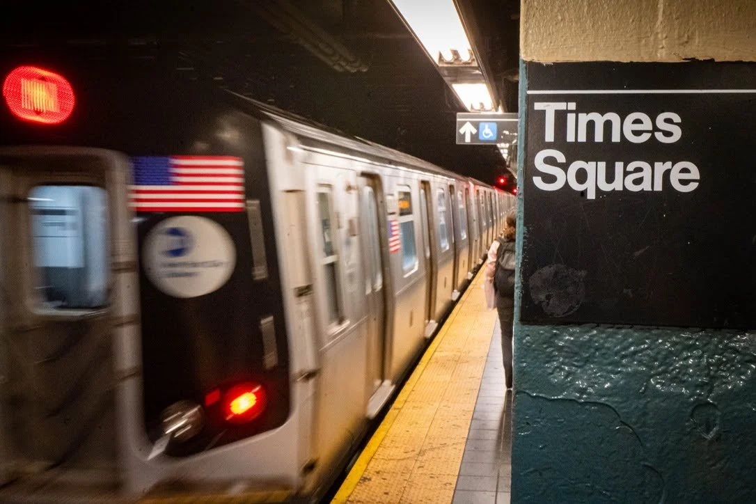 A subway train arriving at Times Square station with a person standing on the platform. The train has American flags and transit symbols on it. The station sign reads 'Times Square'.
