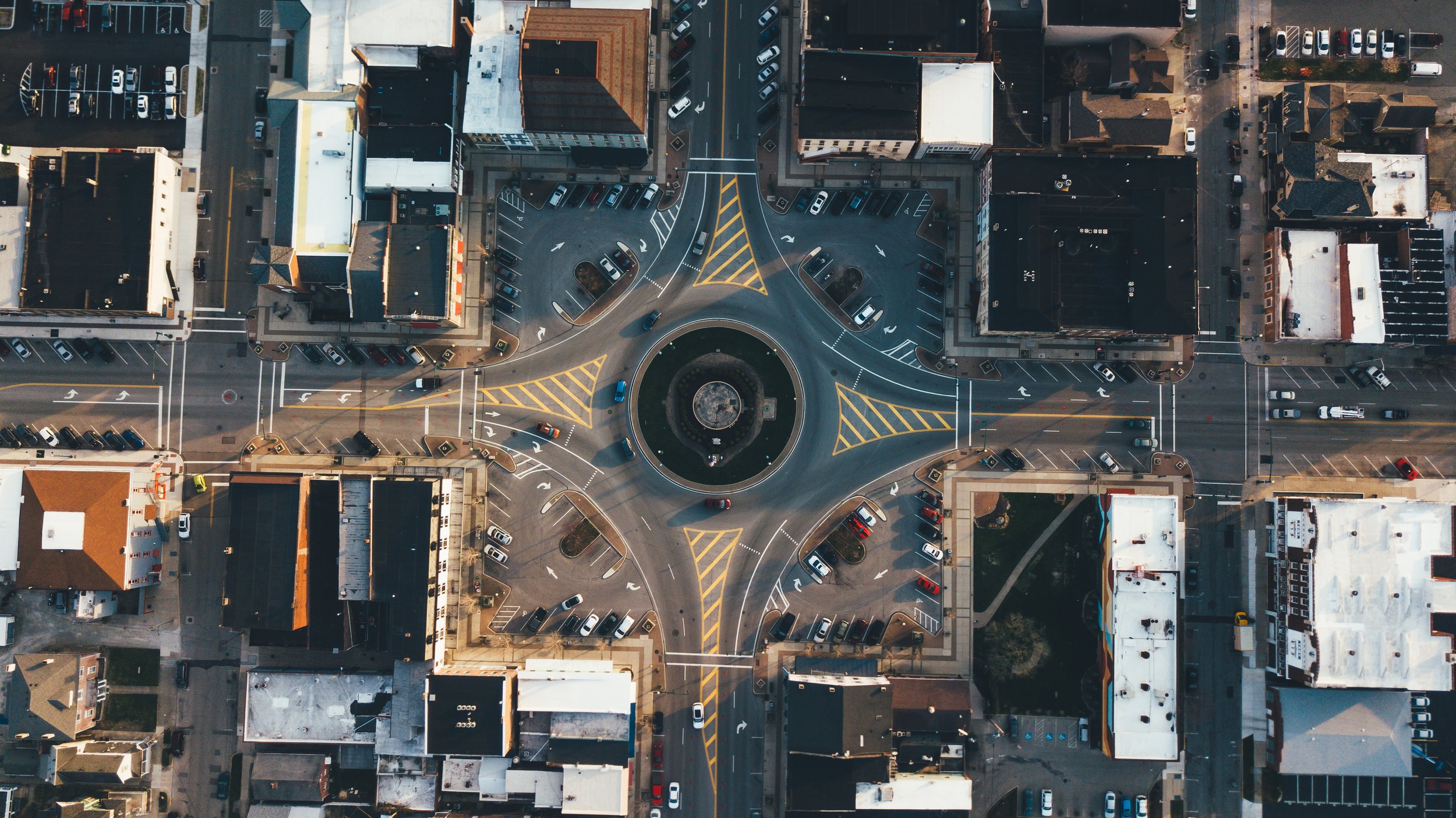 An aerial view of a city intersection with a roundabout, surrounding buildings, and parked cars.