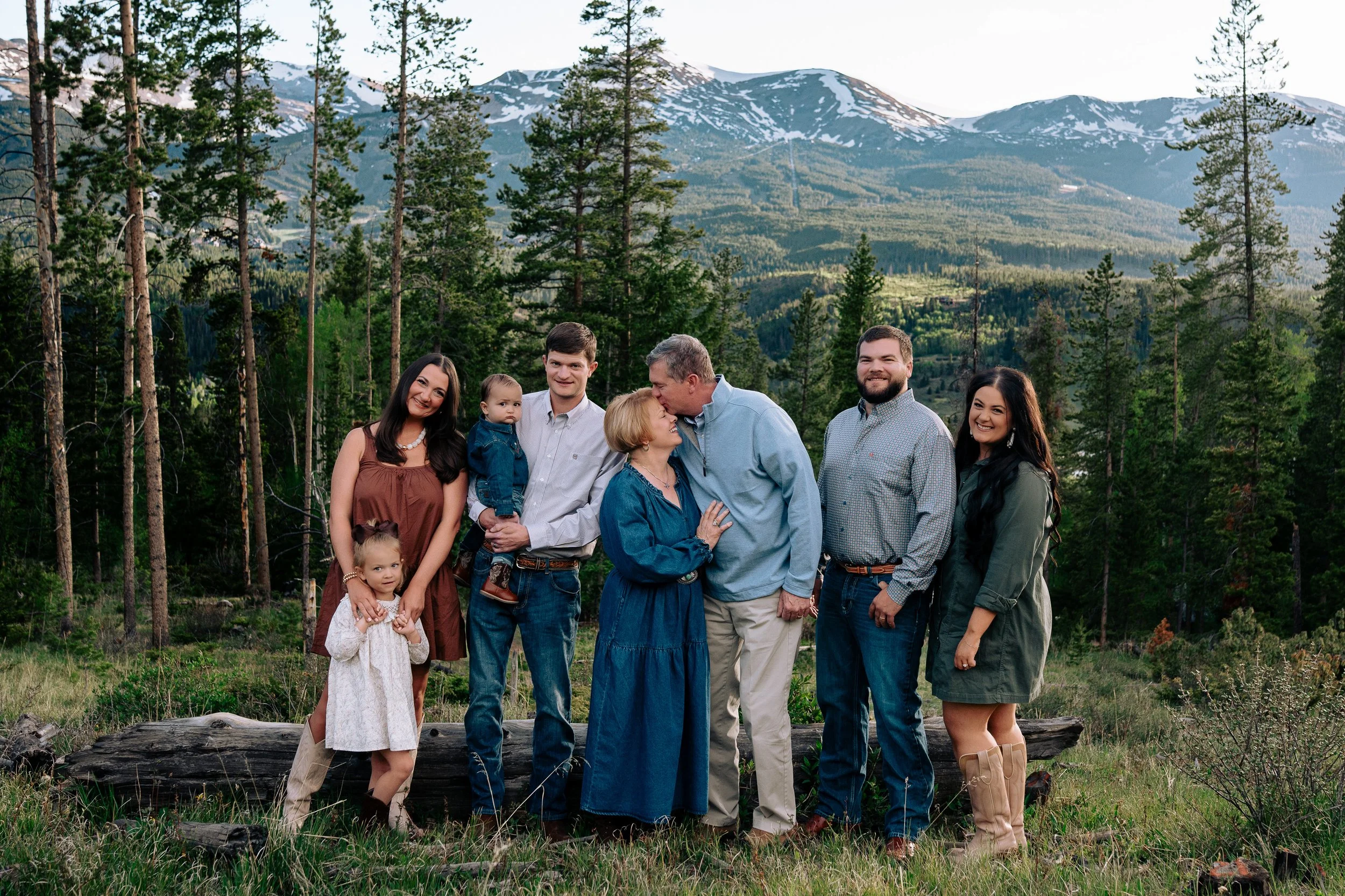 Group of seven people, including three children, standing outdoors in a forested area with mountains in the background. The family is smiling and interacting with each other, dressed in casual clothing, in a scenic natural setting with trees and snow-capped peaks.