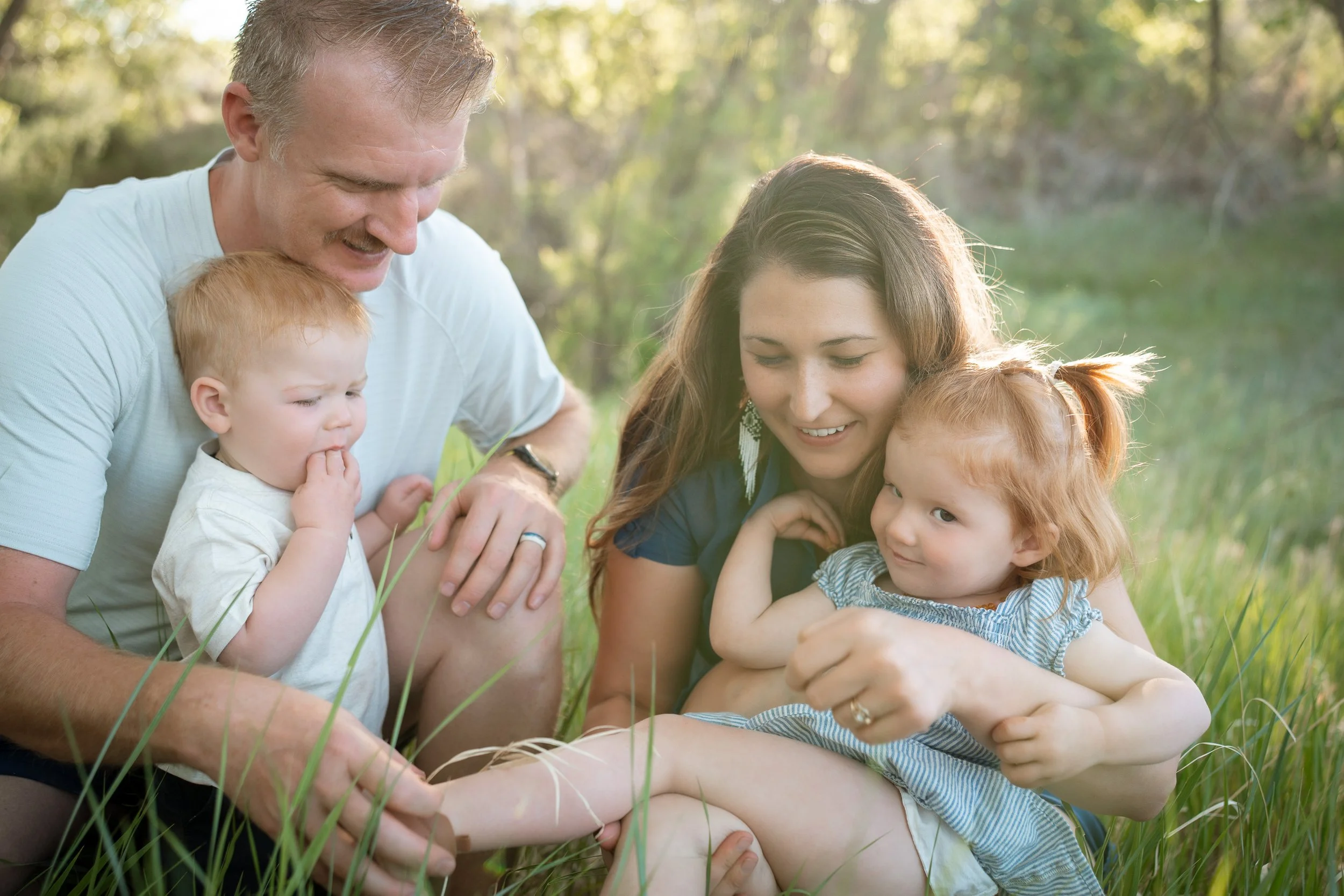 A family of four, including a father, mother, and two young children with red hair, sitting together in a grassy outdoor setting with sunlight filtering through trees, smiling and enjoying quality time.