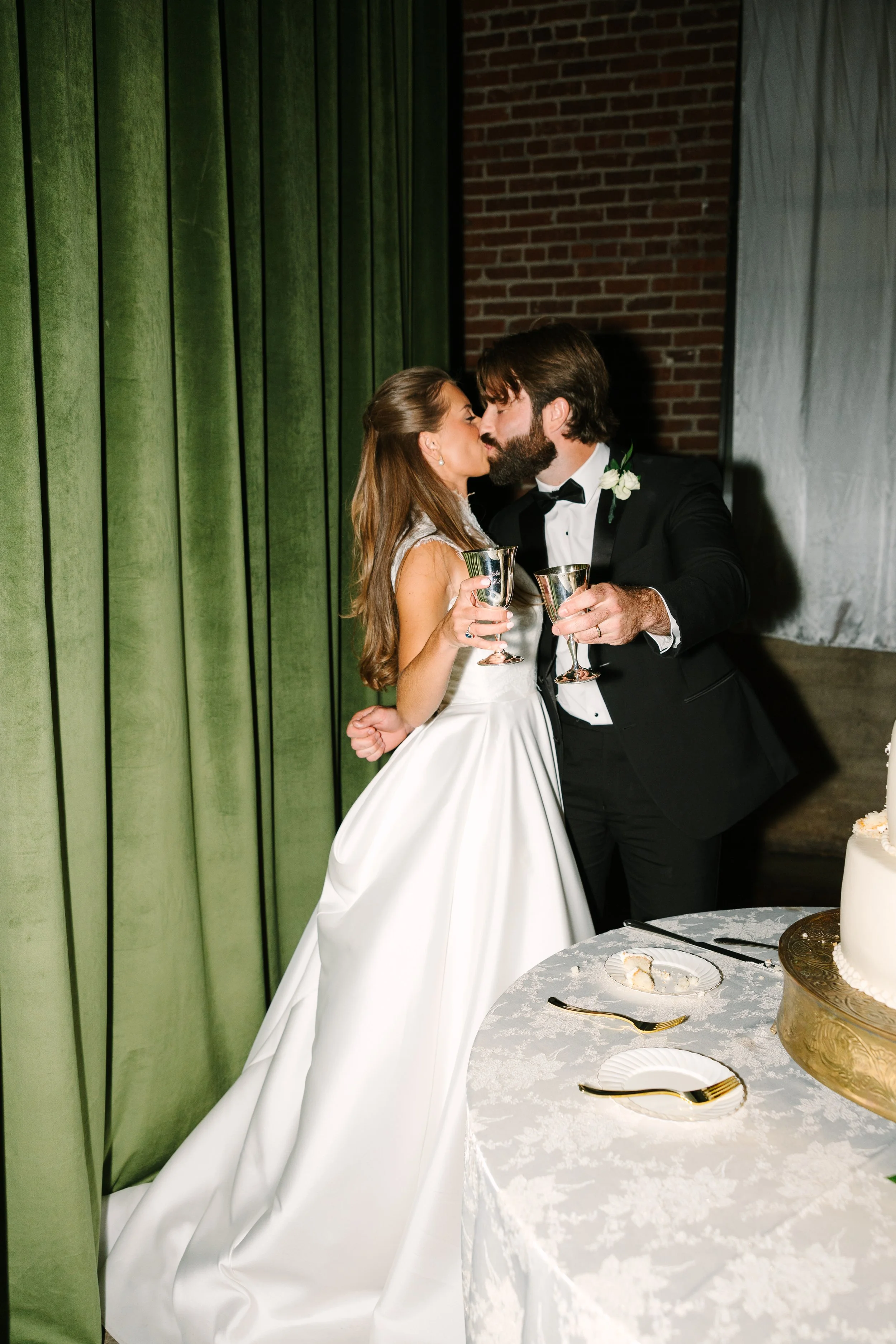 Bride and groom sharing a kiss at their wedding reception, holding glasses of champagne, near a table with wedding cake and decor.