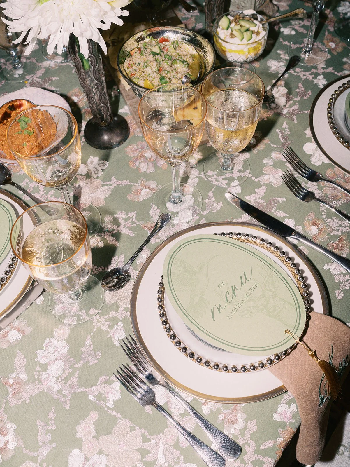 A table setting with wine glasses, a decorative plate with a menu, and various dishes including a salad and bread, arranged on a floral tablecloth with a candle and flower centerpiece.