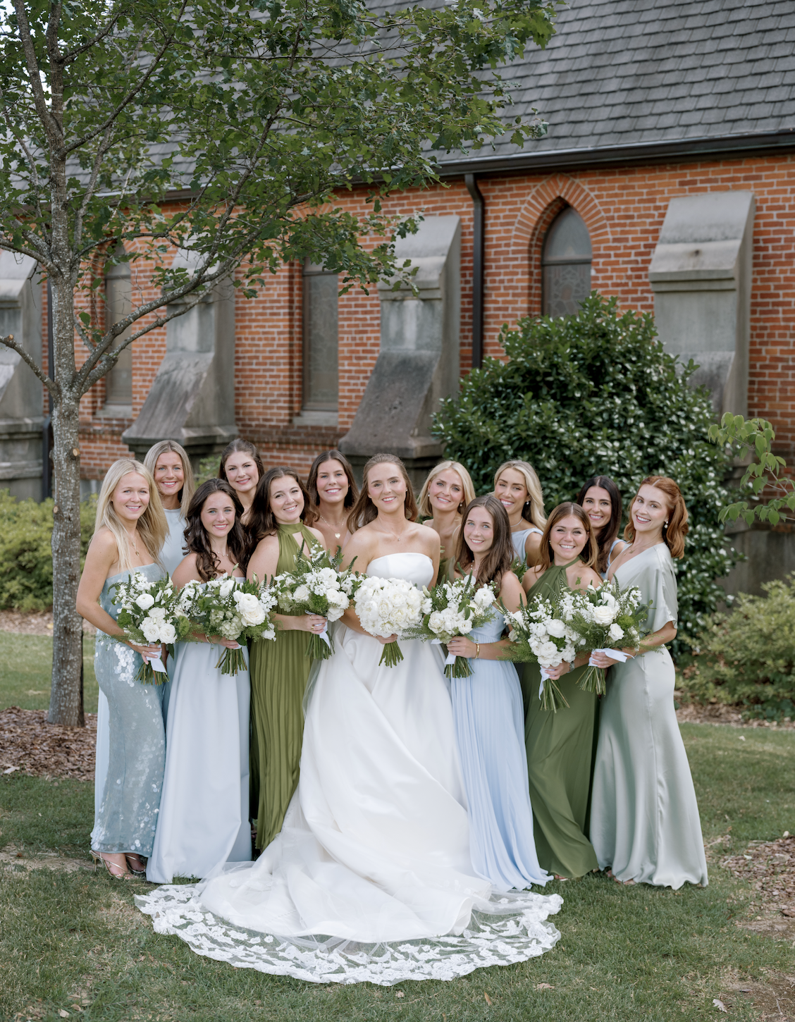 A bride and her bridesmaids with bouquets of white flowers, posed outdoors in front of a brick building with trees and bushes.