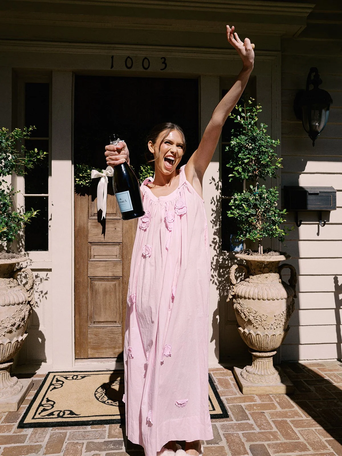 Woman in pink dress celebrating on the front porch of a house, holding a bottle of champagne, with her arm raised.