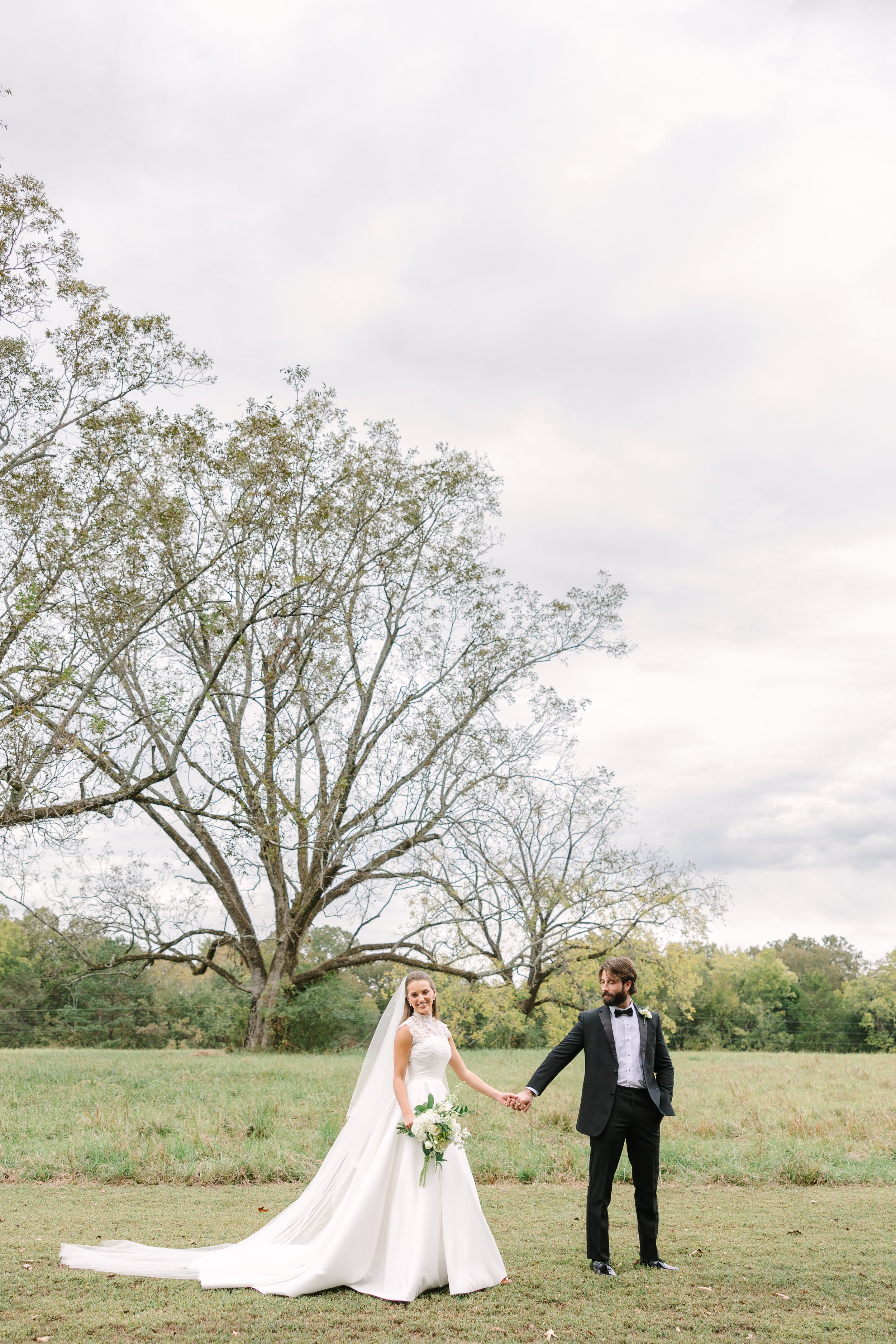 A bride and groom holding hands in a field, with trees and a cloudy sky in the background.