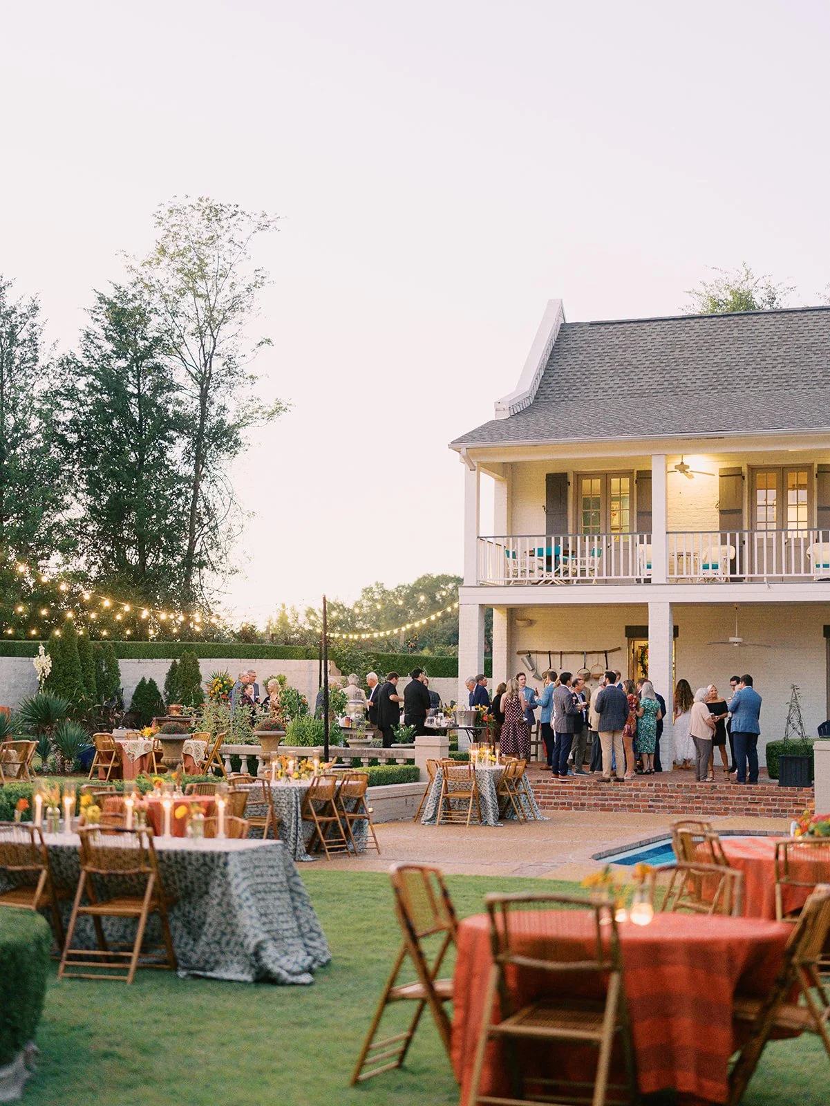 Outdoor evening gathering with people socializing near a house with a balcony, decorated tables with tablecloths and candles, string lights, and a swimming pool.