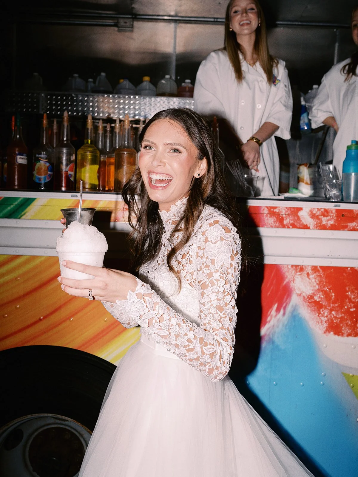 A young woman in a white lace dress holding a large snow cone with a straw, smiling happily at an outdoor event with a colorful trailer behind her and a woman in a white coat standing in the background.