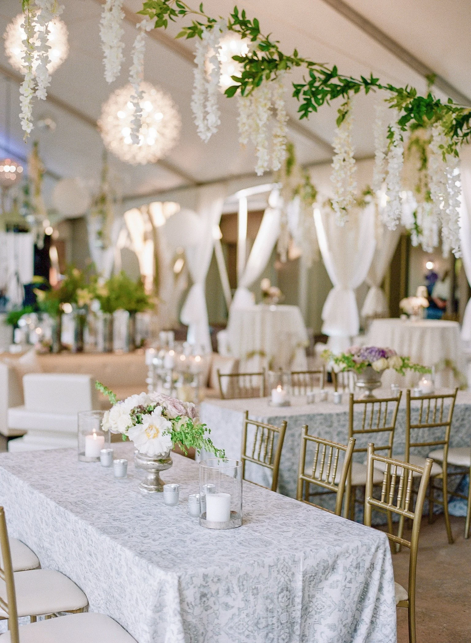 Elegant decorated event space with tables, gold chairs, floral centerpieces, hanging white flowers and candles