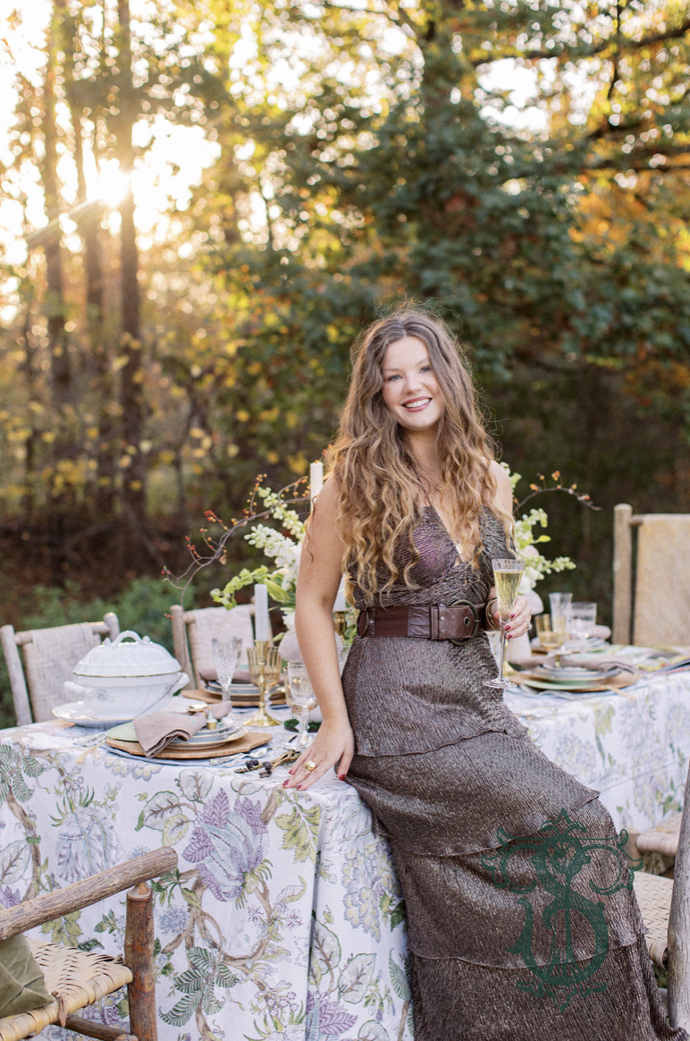 A woman with long, curly hair wearing a dark, strapless dress on an outdoor dining table during sunset in a wooded area.