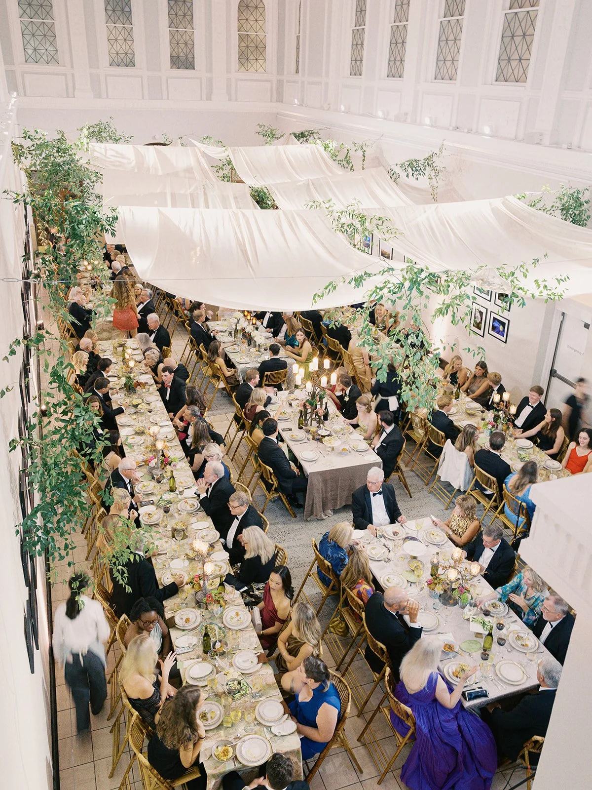 Overhead view of a banquet hall with long rectangular tables set for a formal dinner, decorated with candles, flowers, and tableware. The hall has white walls, large windows, and some greenery hanging from above.