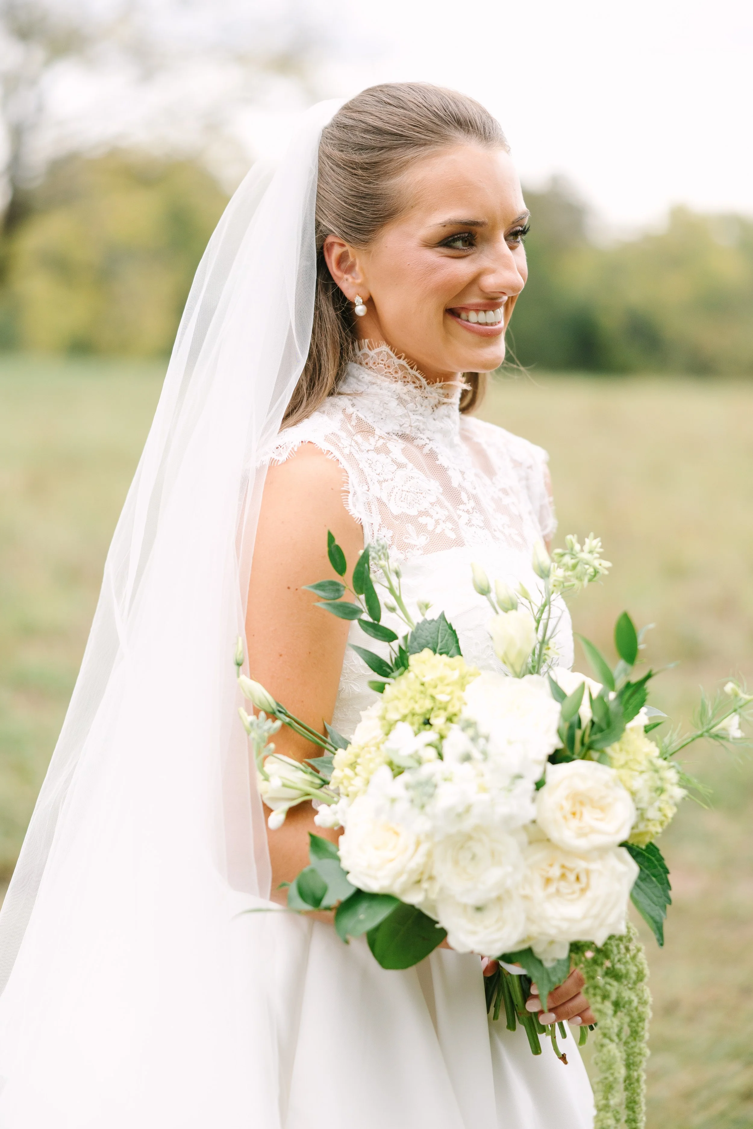 A smiling bride in a lace wedding dress holding a bouquet of white and green flowers outdoors.