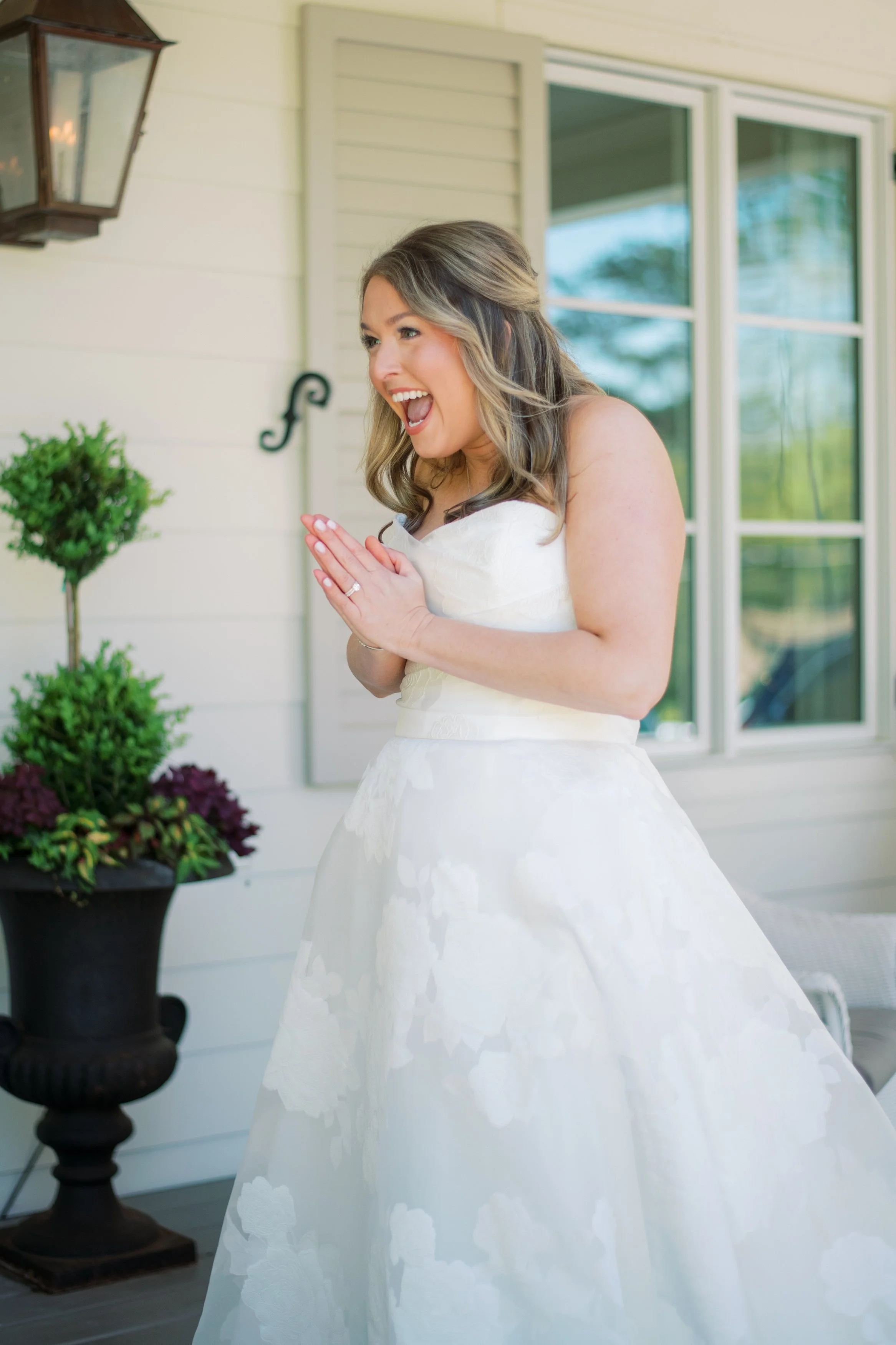 A woman in a wedding dress with a joyful expression on her face, standing outside on a porch with plants and windows in the background.