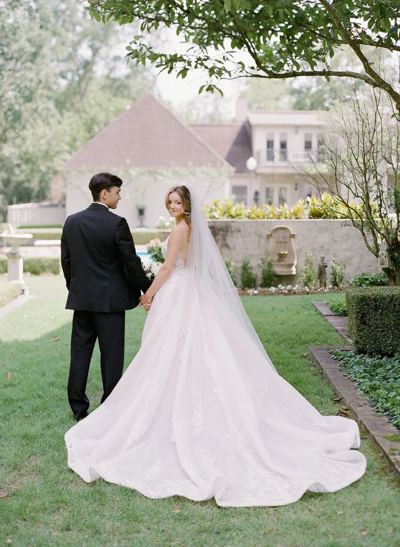 A bride and groom holding hands in a garden, smiling at each other, with a house and greenery in the background.
