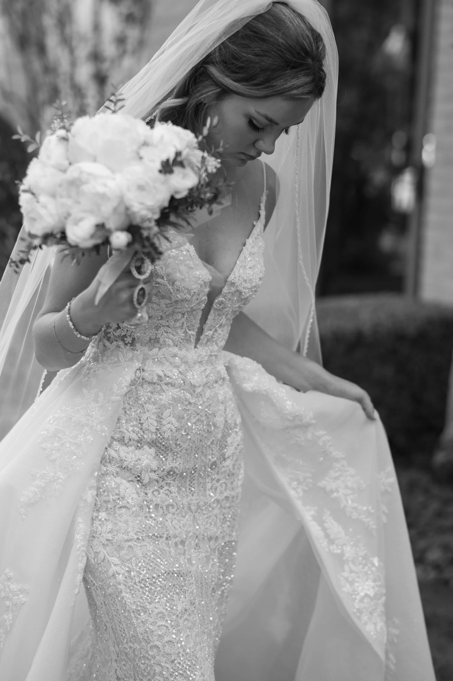 Black and white photo of a bride in a wedding dress holding a bouquet of flowers, looking down, wearing a veil.