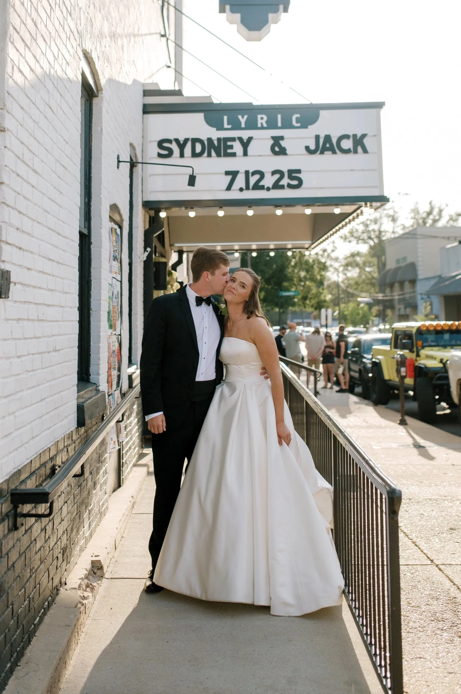 A bride and groom dressed in wedding attire standing outside a theater marquee, which advertises a lyric event featuring Sydney and Jack on July 12th and 25th.