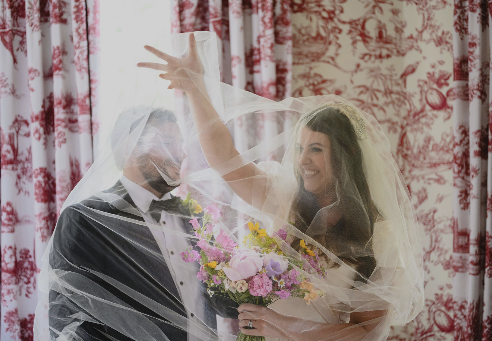 A bride and groom smiling and waving through a sheer veil, with the bride holding a bouquet of colorful flowers in a room with red and white patterned curtains and wallpaper.