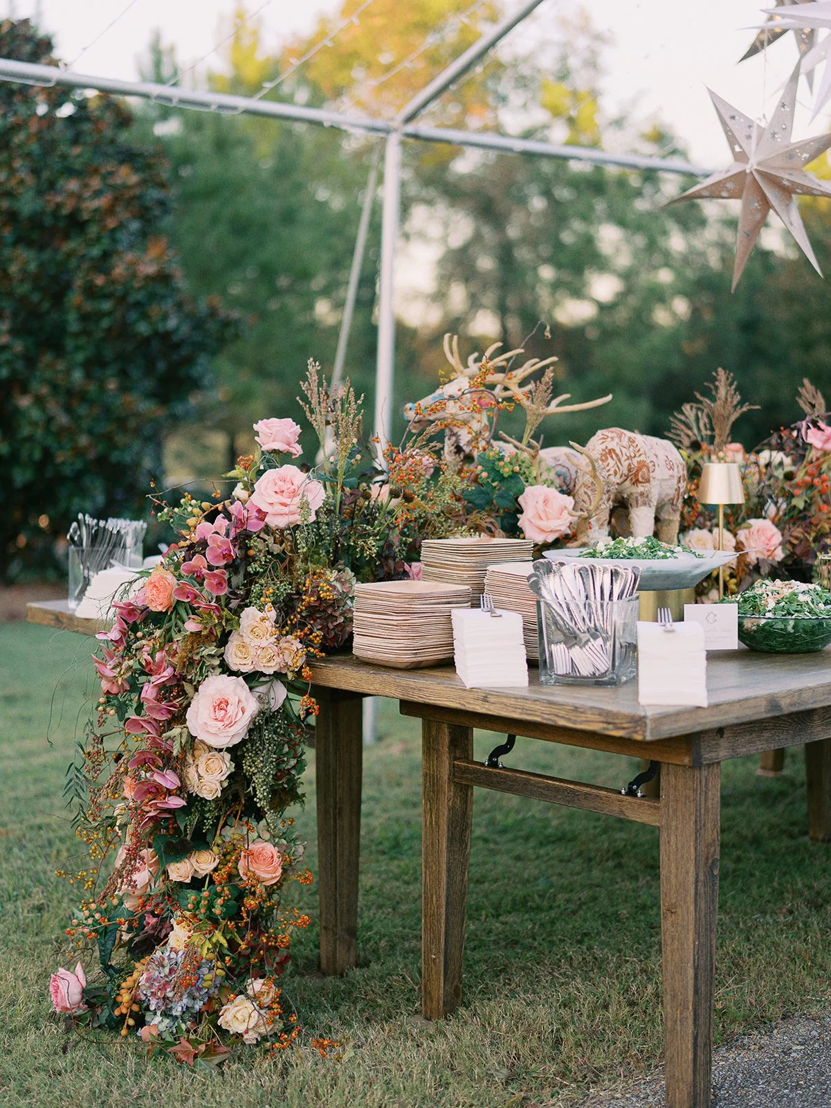 A decorated outdoor table with a floral arrangement, plates, utensils, and decorative objects including star-shaped ornaments and an elephant sculpture, set against greenery and trees.