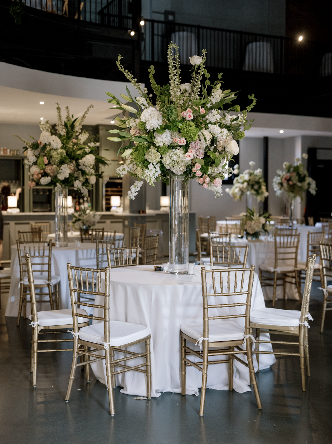 A large round table decorated with a tall glass vase filled with white and pink flowers, including roses and hydrangeas, in an event hall set for a celebration or wedding reception.