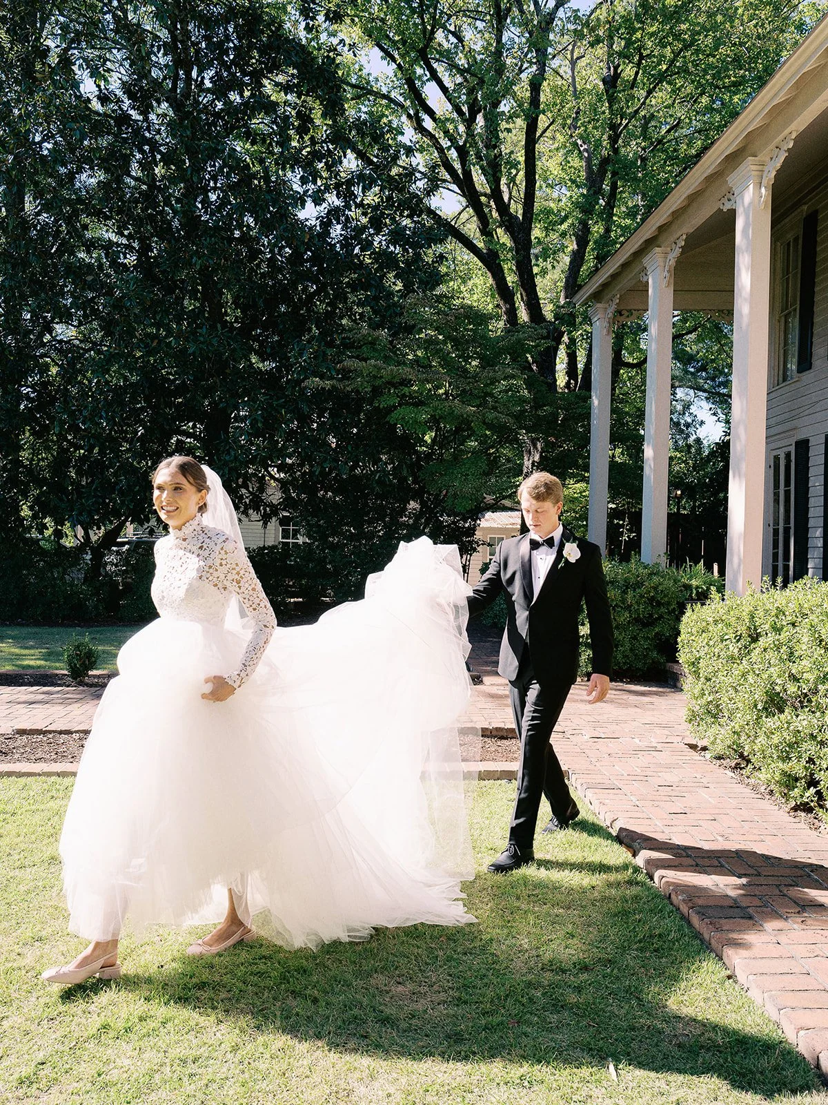 A bride and groom walking outside on a sunny day, with the bride smiling and holding her dress, the groom in a black tuxedo, near a white house with columns, green trees, and bushes in the background.