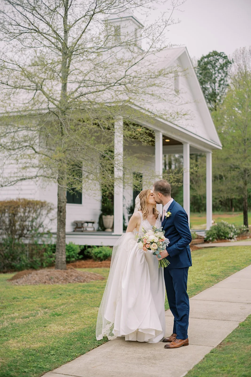 Bride and groom sharing a kiss outdoors on their wedding day outside a white house with a porch, trees, and greenery.
