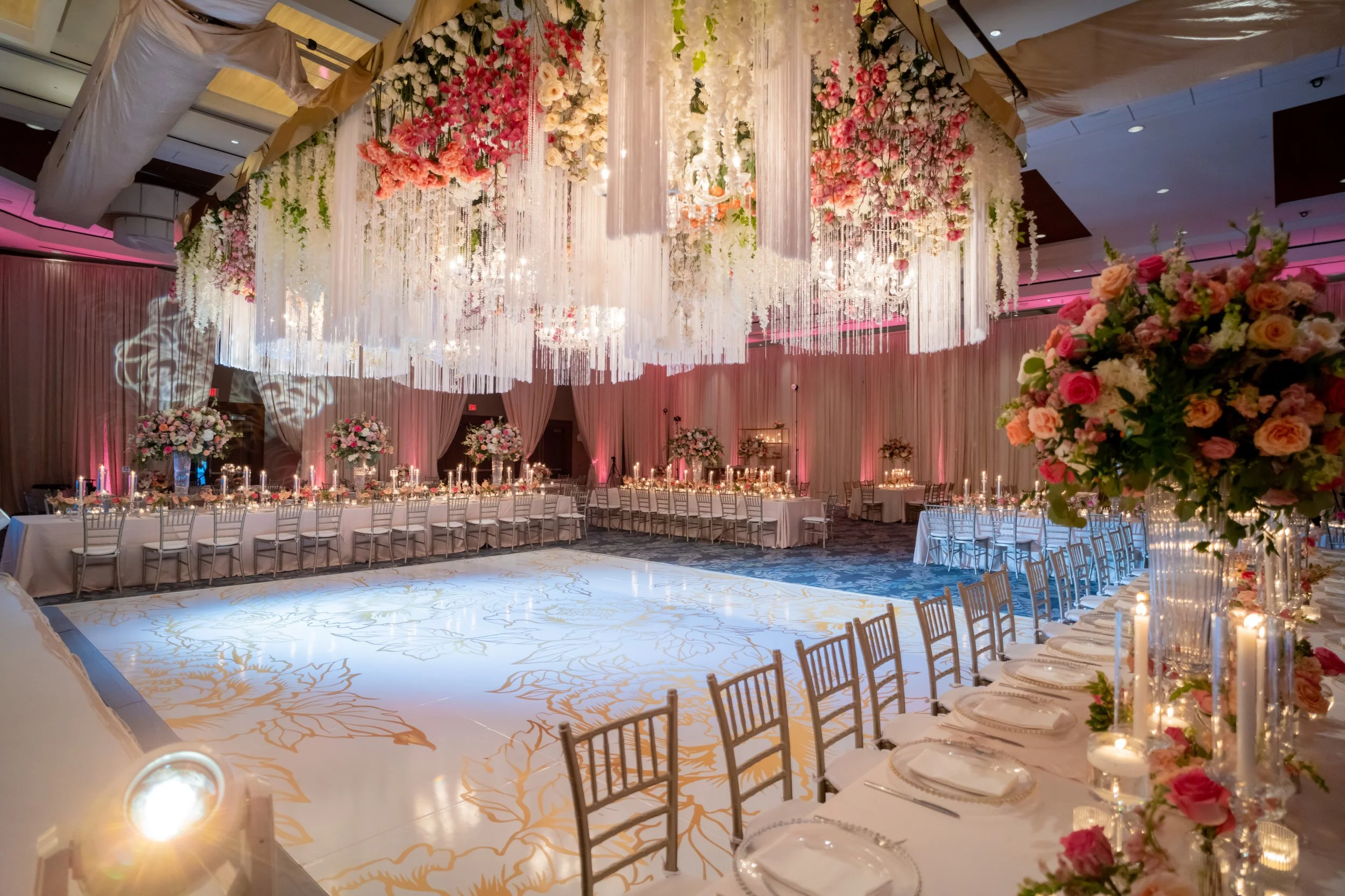 Elegant wedding reception hall decorated with large floral arrangements, hanging floral installations, and candlelit tables under a floating floral chandelier.