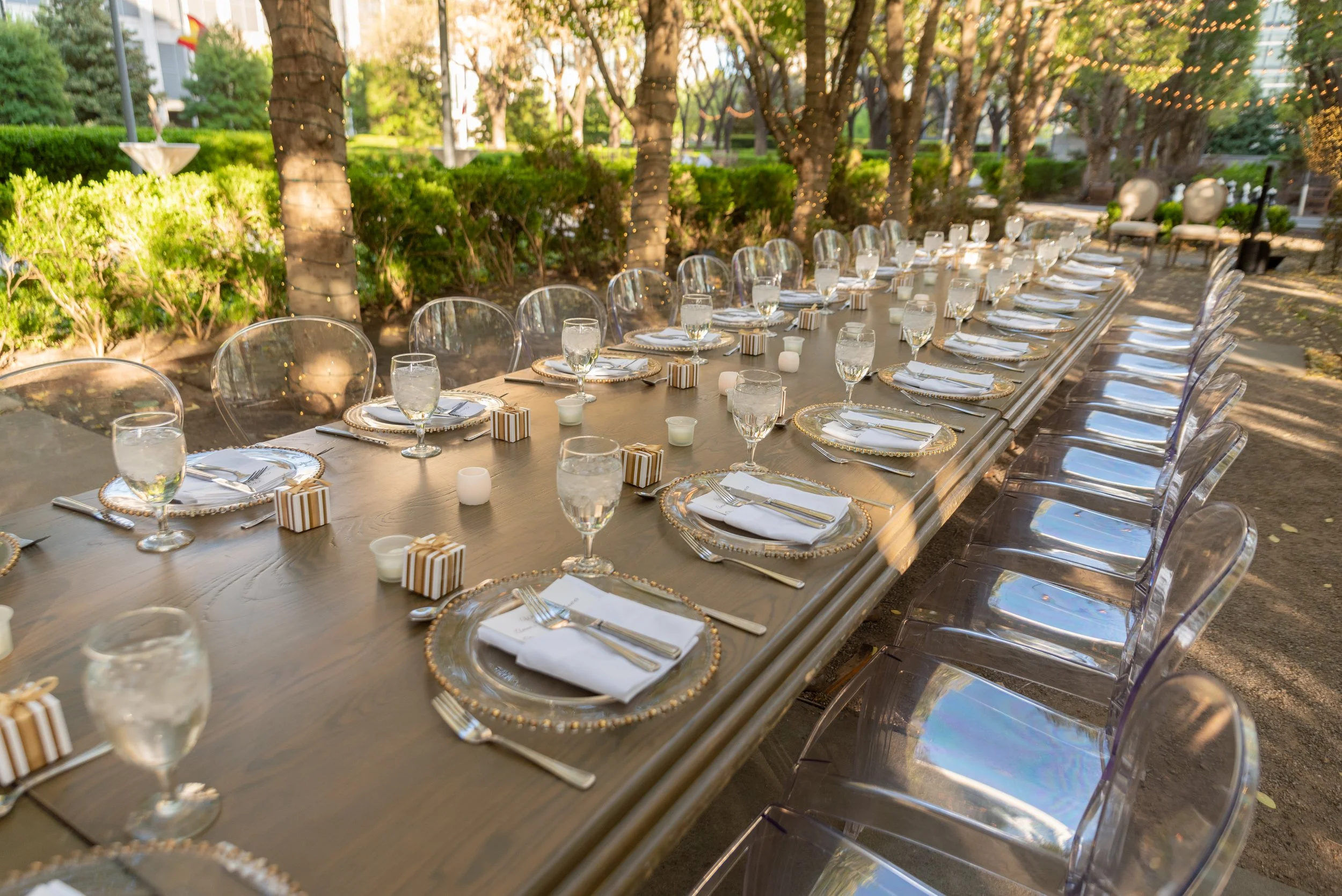 Long outdoor dining table set for a celebration with gold-rimmed plates, cloth napkins, glasses of water, small gift boxes, and candles, surrounded by transparent chairs, in a garden with string lights and shaded trees.