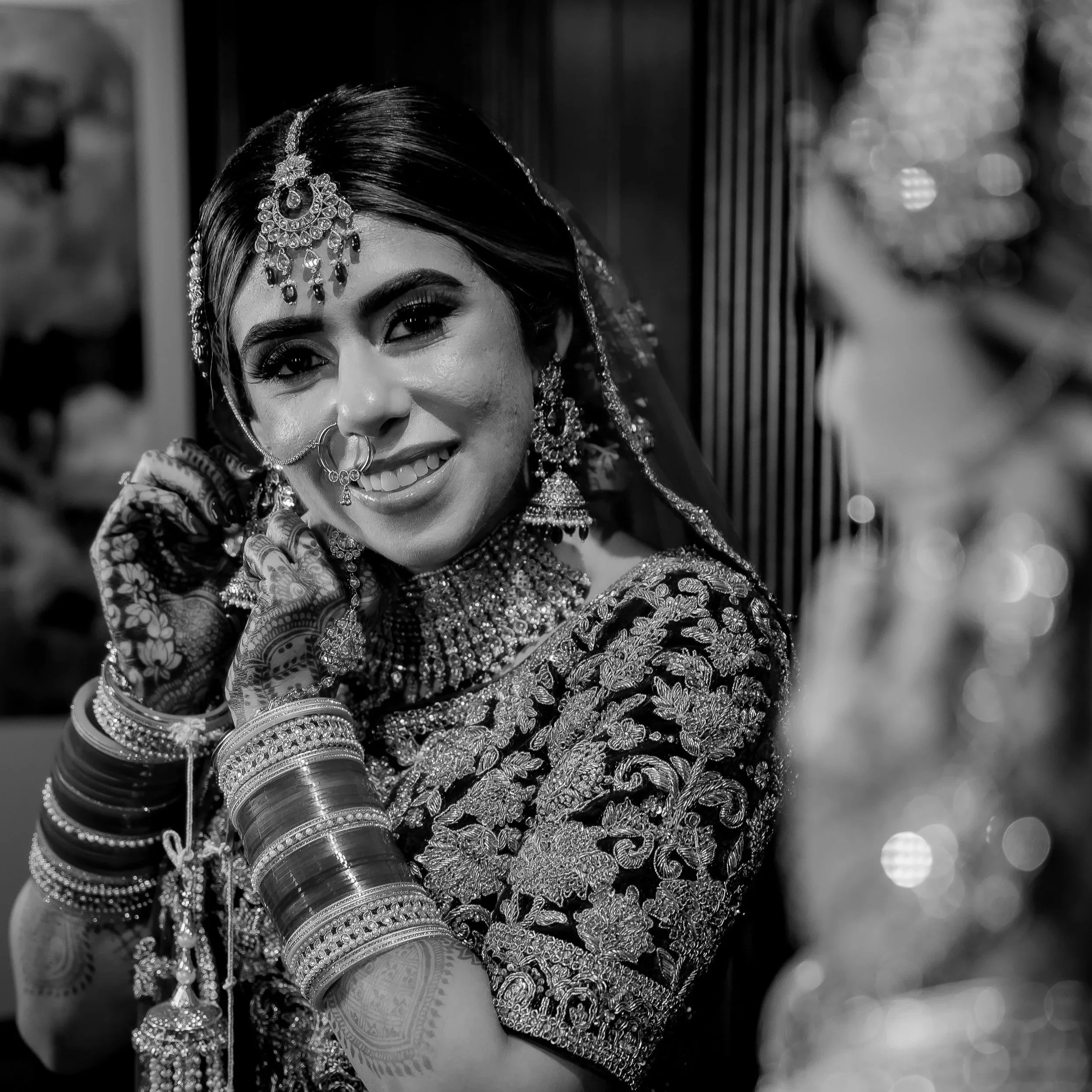 A smiling woman dressed in traditional Indian bridal attire, wearing ornate jewelry and bangles, adjusting her earring.