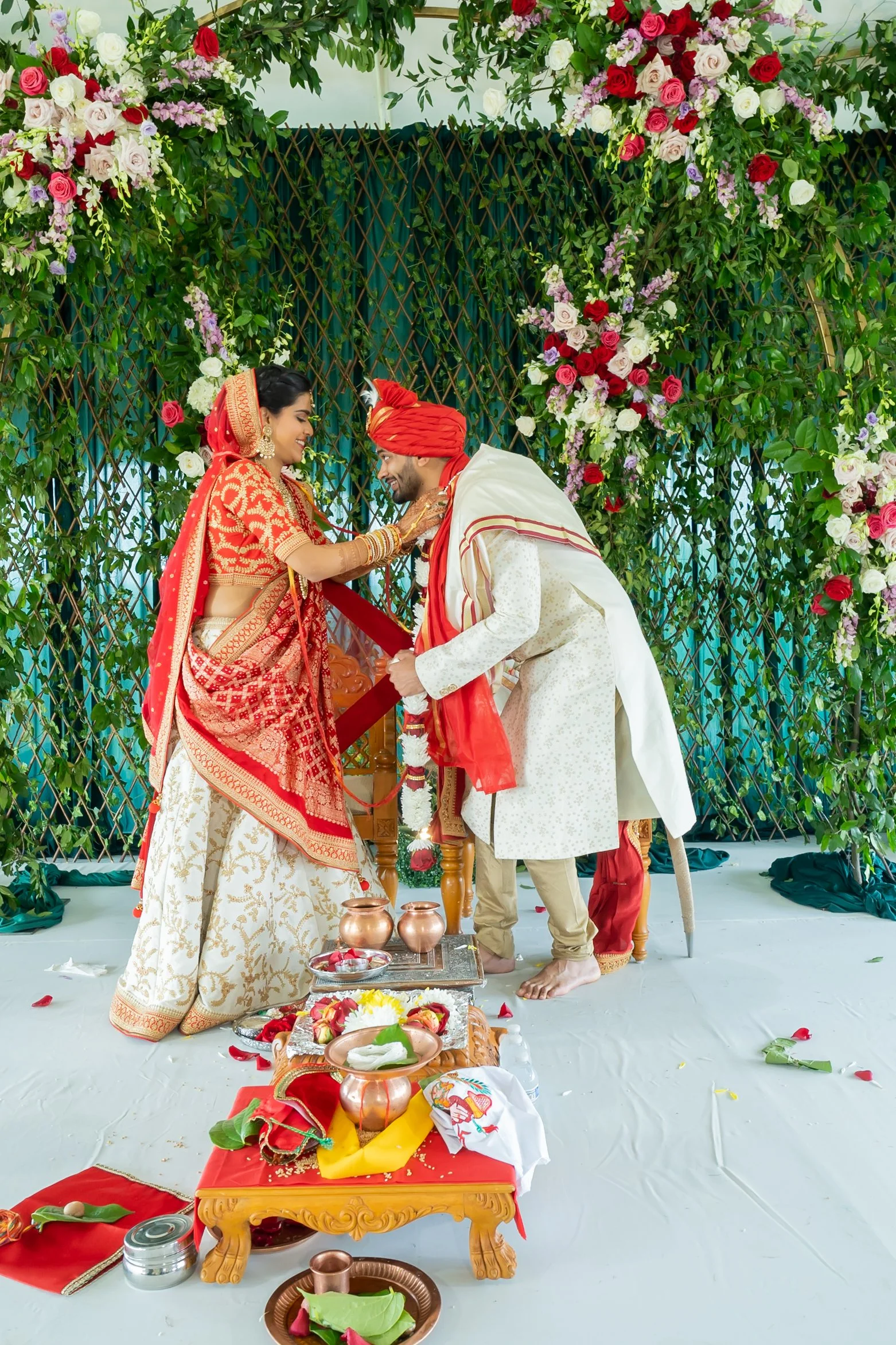 Indian bride and groom during a wedding ceremony, standing on a decorated stage with flowers and traditional ceremonial items in front of them.