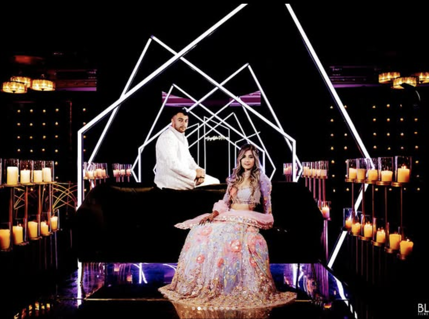 A man in a white shirt and a woman in a pastel pink and purple gown sitting in a modern, geometric light installation with candles on either side.