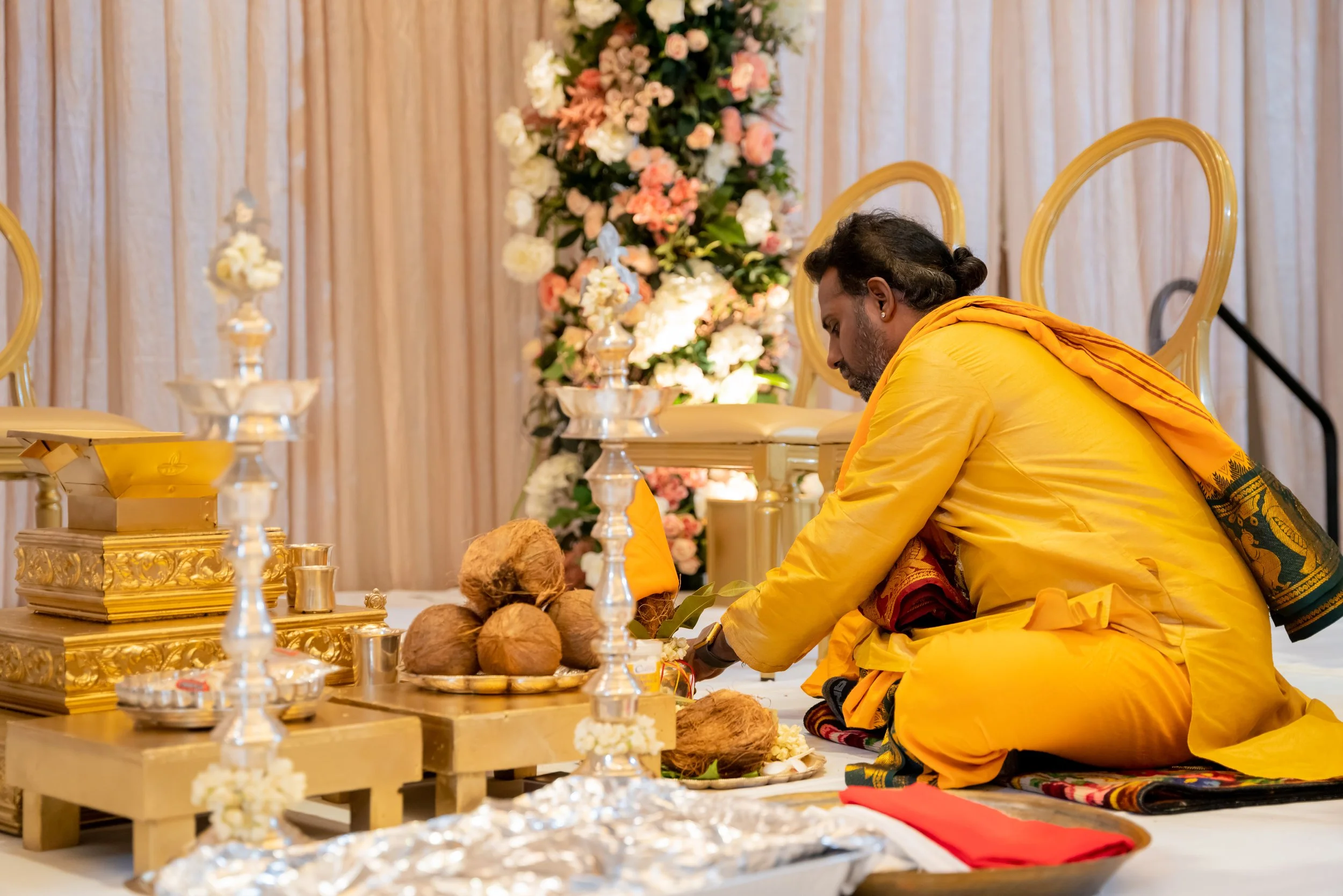 A man dressed in yellow traditional attire kneeling on the floor during a religious ceremony, with coconut and other ritual items in front of him, decorated with flowers in the background.