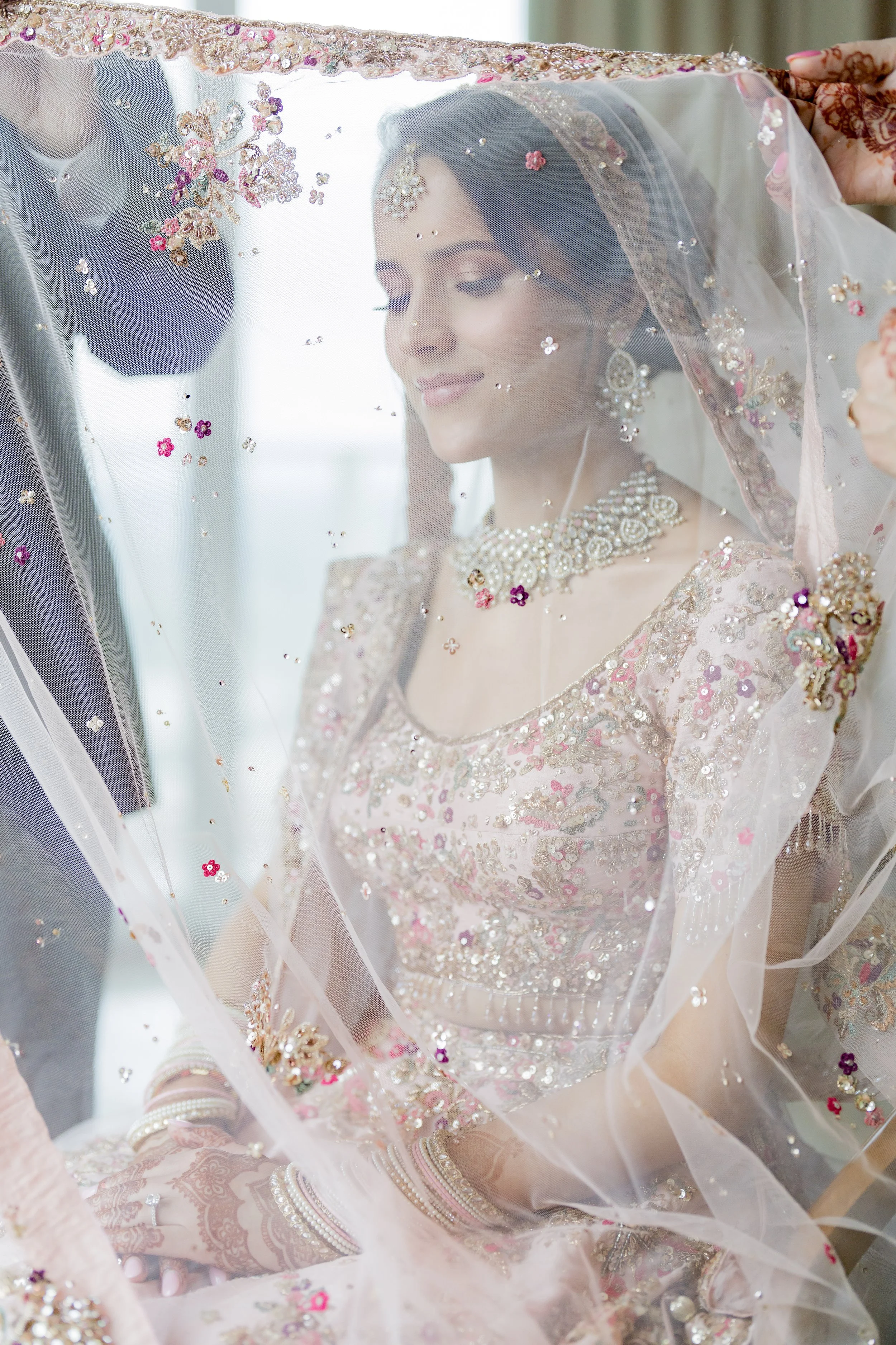 A bride dressed in traditional Indian wedding attire, adorned with jewelry, sitting with a gentle smile behind a sheer veil with colorful embroidery and embellishments.