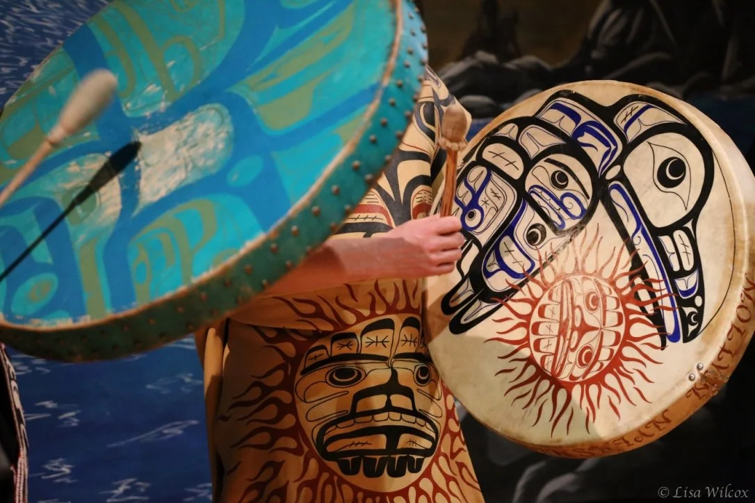 Two performers playing large drums decorated with traditional Indigenous art, featuring animal faces and intricate patterns.